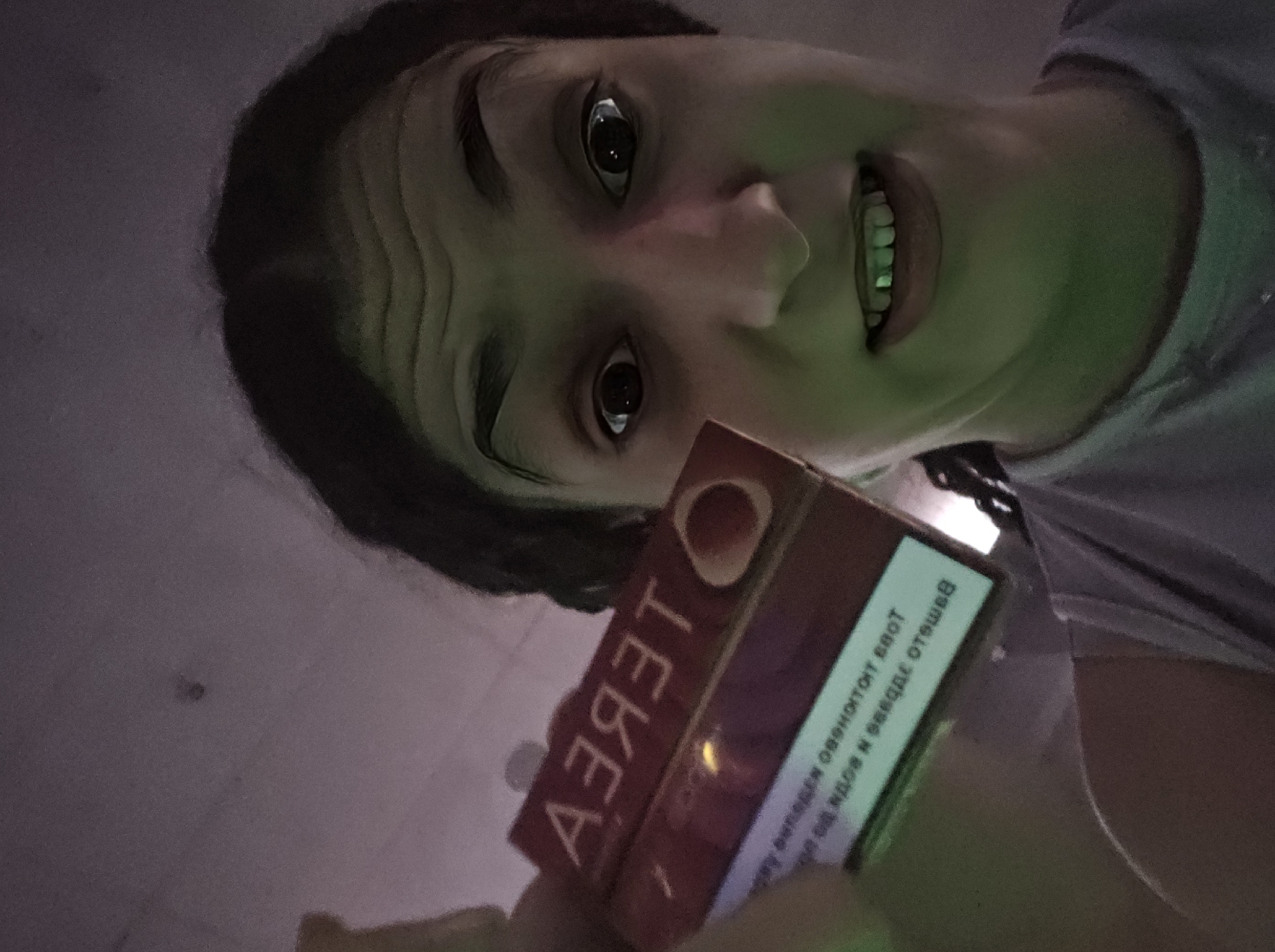 A person holds a pack of "TEREA" brand tobacco cigarettes. The text on the side of the pack is in Bulgarian. The lighting casts a green hue on the scene. The person is likely indoors with a white wall in the background. Pleven, Bulgaria, is not discernable from the details in the image.
