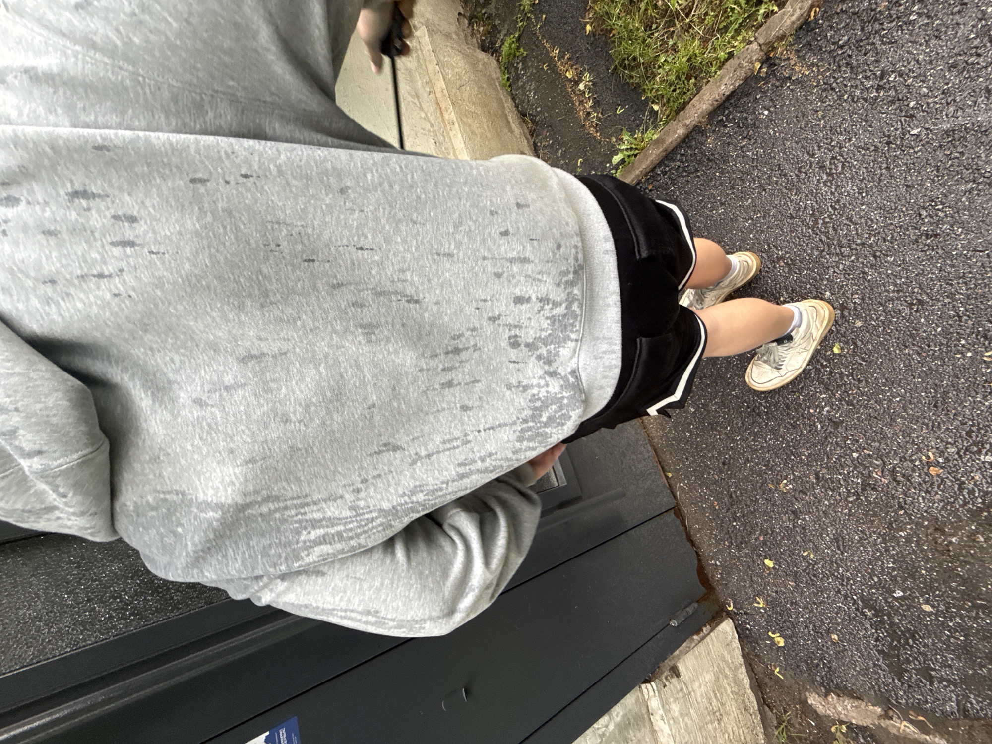 The image shows the back of a person wearing a grey hooded sweatshirt and black shorts with white trim. The sweatshirt appears wet, with dark streaks indicating water stains. The person is standing on a wet, dark asphalt surface, possibly a sidewalk or path, with scattered leaves and debris. To the left of the person is a dark, possibly metal, door or wall. To the right, there is overgrown grass and a concrete curb or edge. The overall impression is that of an outdoor environment, possibly in damp or rainy weather. The specific location in Saint Petersburg, Russia cannot be determined from the visual information in the image.