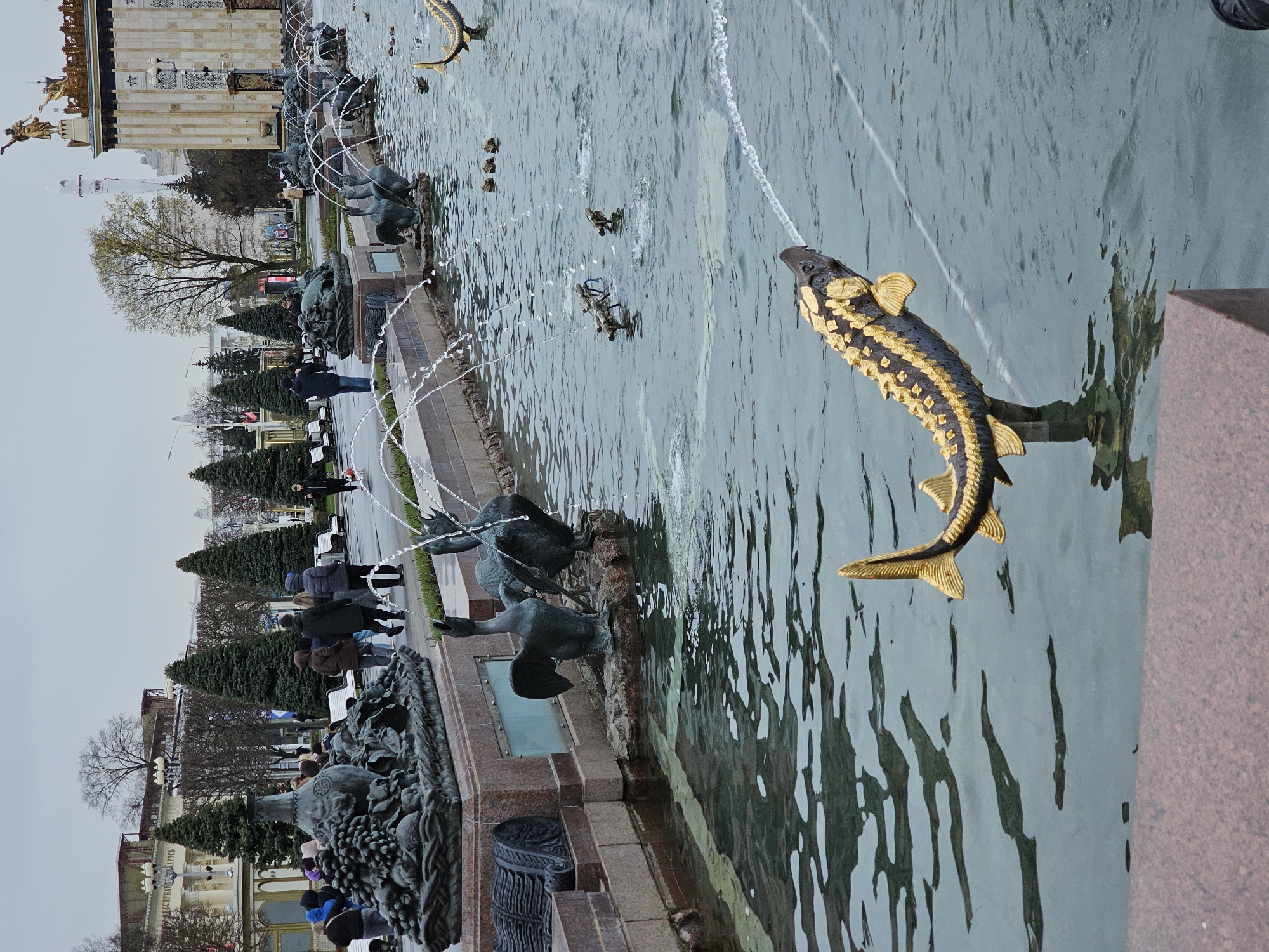 The image captures a section of the Friendship of Peoples Fountain in Moscow, Russia. The fountain features several bronze sturgeon sculptures spouting water. People are visible along the edge of the fountain, seemingly observing the display. The surrounding environment includes trees, buildings, and architectural details typical of Moscow.