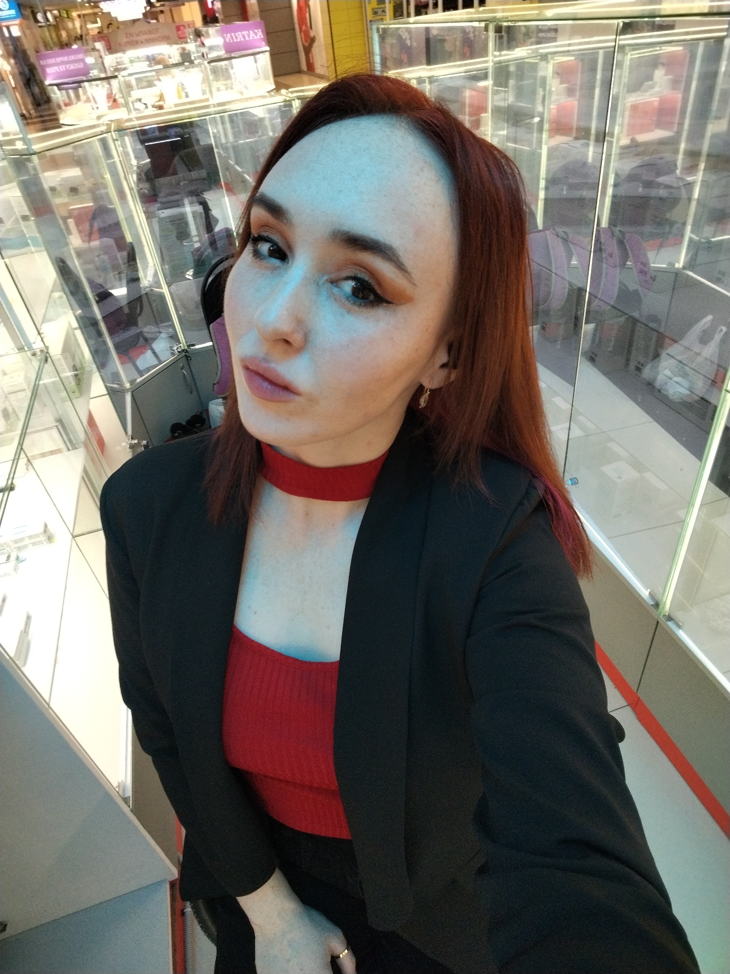 A woman with long, reddish-brown hair poses for a selfie, looking directly into the camera with a confident expression. She has noticeable eye makeup and is wearing a red, ribbed top with a matching red choker, under a black blazer. A gold-colored ring is visible on her right hand.
The scene is an indoor commercial environment, likely a shopping mall or a large retail store in Izhevsk, Russia. The background is dominated by numerous glass display cases or railings, creating reflections of other parts of the building, including shelves and merchandise. The glass also distorts and reverses text on promotional signs. Clearly visible reversed text includes "КАТЯ" (Katya) and "АКЦИЯ" (Aktsiya, meaning "Promotion" or "Sale"). Another partially visible and reversed inscription reads "EN MBABOT," likely part of "В РАБОТЕ" (V Rabote, meaning "In Operation" or "Working"). The lighting is bright and consistent, typical of an indoor commercial setting during the day.