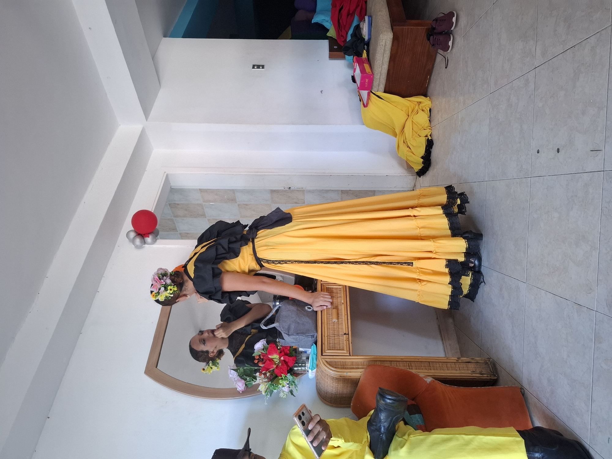 The image captures an indoor scene, seemingly a residence in Playa El Agua, Venezuela. A woman dressed in a long, yellow dress with black trim is the focal point, captured bending slightly toward a circular mirror. She has a floral headpiece. Her reflection is visible in the mirror. A man wearing a yellow shirt is seated, partly out of frame, in what seems to be a wicker chair, using a smartphone. The room's decor is basic; a tiled floor leads up to white walls. There are random items and clutter visible, including clothing and a shoe rack with footwear. A red balloon and other decorations are placed near a doorway in the background. The overall impression is of a casual, indoor setting, likely during or after preparations for a festivity.