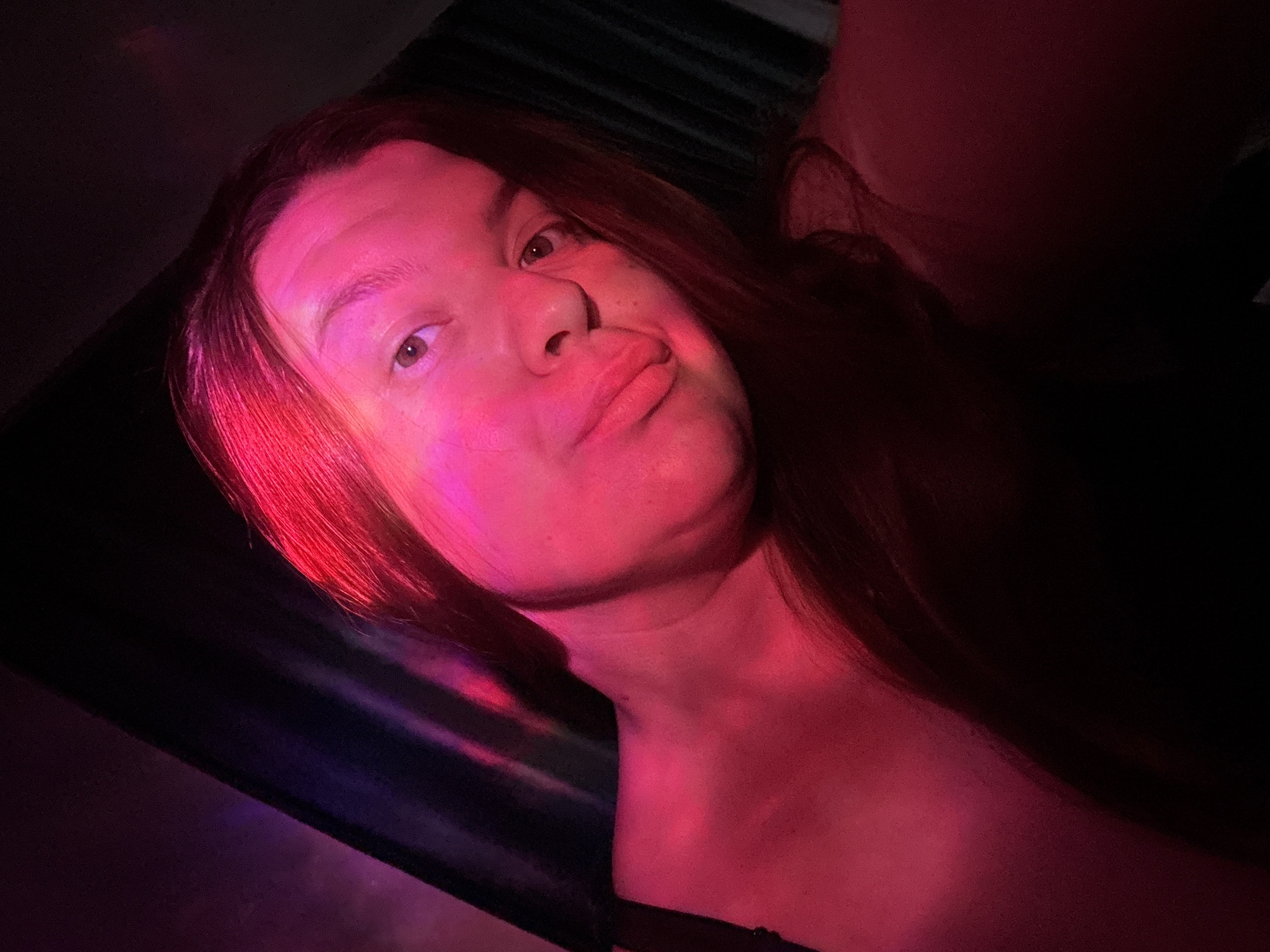 A medium close-up shot of a person's face and upper torso, angled upwards. The individual has light skin, brown hair, and is illuminated with red and purple lighting, creating dramatic shadows and highlights. The person is pursing their lips in a slight pout. A dark, shiny surface is visible on the left, possibly a part of a seat or wall. A dark shape is visible near the right shoulder, possibly the strap of a garment. The location is Марковский, Russia.