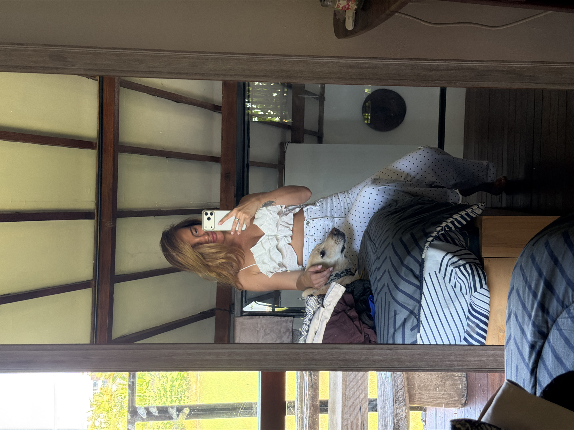 A woman with blonde hair, wearing a white ruffled crop top and white polka-dot pants, is taking a mirror selfie in a room. She is holding a smartphone in her right hand. A light-colored dog is resting its head on a bed next to her. The room appears to be a bedroom with a bed covered in patterned sheets and blankets, and what looks like a wooden ceiling. In the reflection, a large window shows a green, leafy outdoor environment. The mirror is framed by a wooden border, and the overall scene suggests a casual, indoor setting in North Kuta, Indonesia.