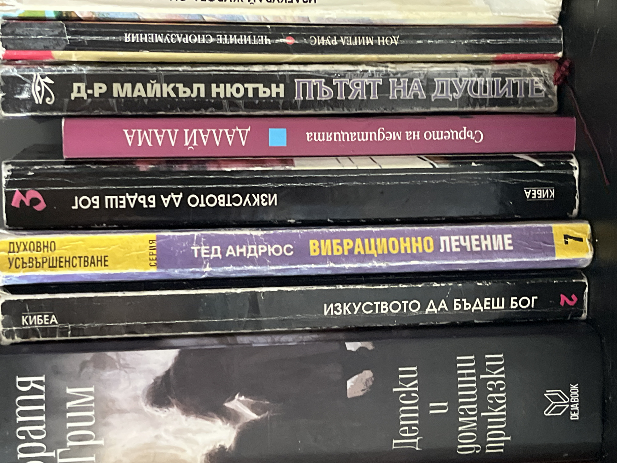 A vertical shot displays a stack of eight books on a dark surface, likely a shelf or table. The books vary in spine color, predominantly black, with some pink, purple, and yellow. Several books are stacked upside down, indicating a casual arrangement.
From top to bottom, the visible titles and authors are:
1. A partially visible book with a light-colored spine.
2. "ЧЕТИРИТЕ СПОРАЗУМЕНИЯ" (The Four Agreements) by ДОН МИГЕЛ РУИС (Don Miguel Ruiz).
3. "ПЪТЯТ НА ДУШИТЕ" (Journey of Souls) by Д-Р МАЙКЪЛ НЮТЪН (Dr. Michael Newton).
4. "Сърцето на медитацията" (The Heart of Meditation) by ДАЛАЙ ЛАМА (Dalai Lama), with a pink spine and a small blue square, stacked upside down.
5. "ИЗКУСТВОТО ДА БЪДЕШ БОГ" (The Art of Being God), published by КИБЕА (KIBEA), stacked upside down.
6. "ВИБРАЦИОННО ЛЕЧЕНИЕ" (Vibrational Healing) by ТЕД АНДРЮС (Ted Andrews), part of the "ДУХОВНО УСЪВЪРШЕНСТВАНЕ" (Spiritual Self-Improvement) series, volume 7, featuring a purple and yellow spine.
7. "ИЗКУСТВОТО ДА БЪДЕШ БОГ" (The Art of Being God), published by КИБЕА (KIBEA), volume 2.
8. "Детски и домашни приказки" (Children's and Household Tales) by Братя Грим (Brothers Grimm), published by DEJA BOOK, stacked upside down.
The collection includes spiritual and philosophical works alongside classic children's literature.