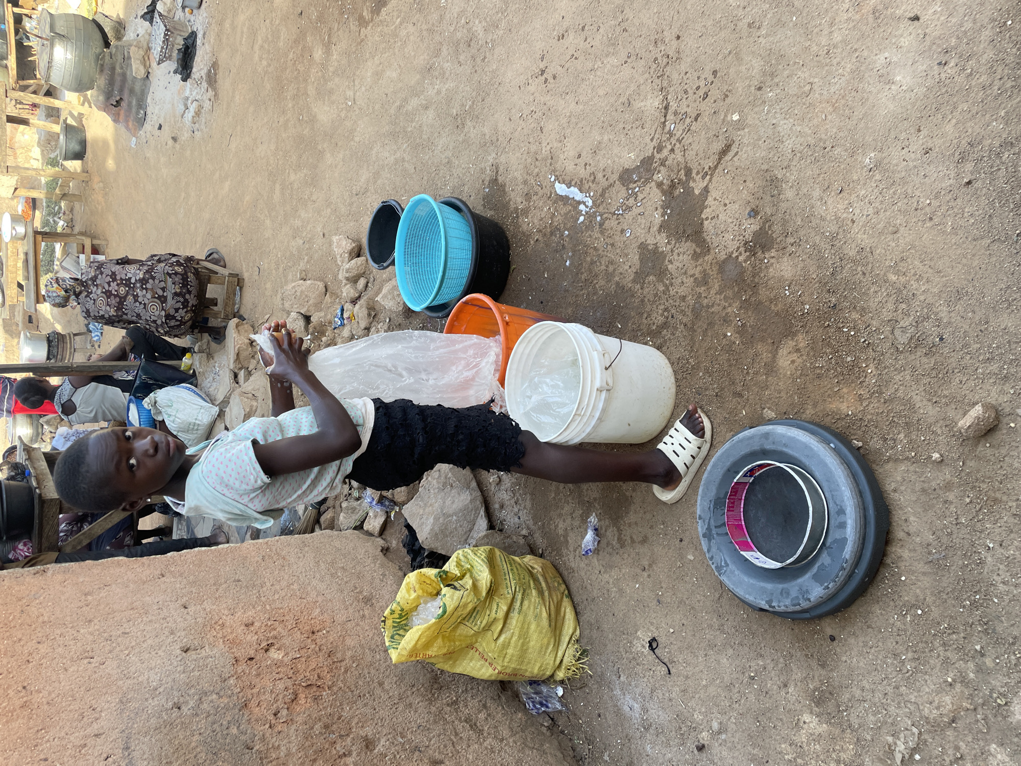The image shows a young person of African descent lying on their side in what appears to be an outdoor market in Jos, Nigeria. The ground is dirt and concrete with scattered debris. The person is wearing a light-colored patterned shirt, black shorts, and white sandals. They are holding something in their hands that is wrapped in a clear plastic bag. Surrounding the person are several plastic buckets of various colors, a yellow sack, and a large grey container with a smaller black and red pan inside. In the background, there are structures made of wood and metal, and other people are visible. The scene suggests a bustling outdoor environment with market-related activities.