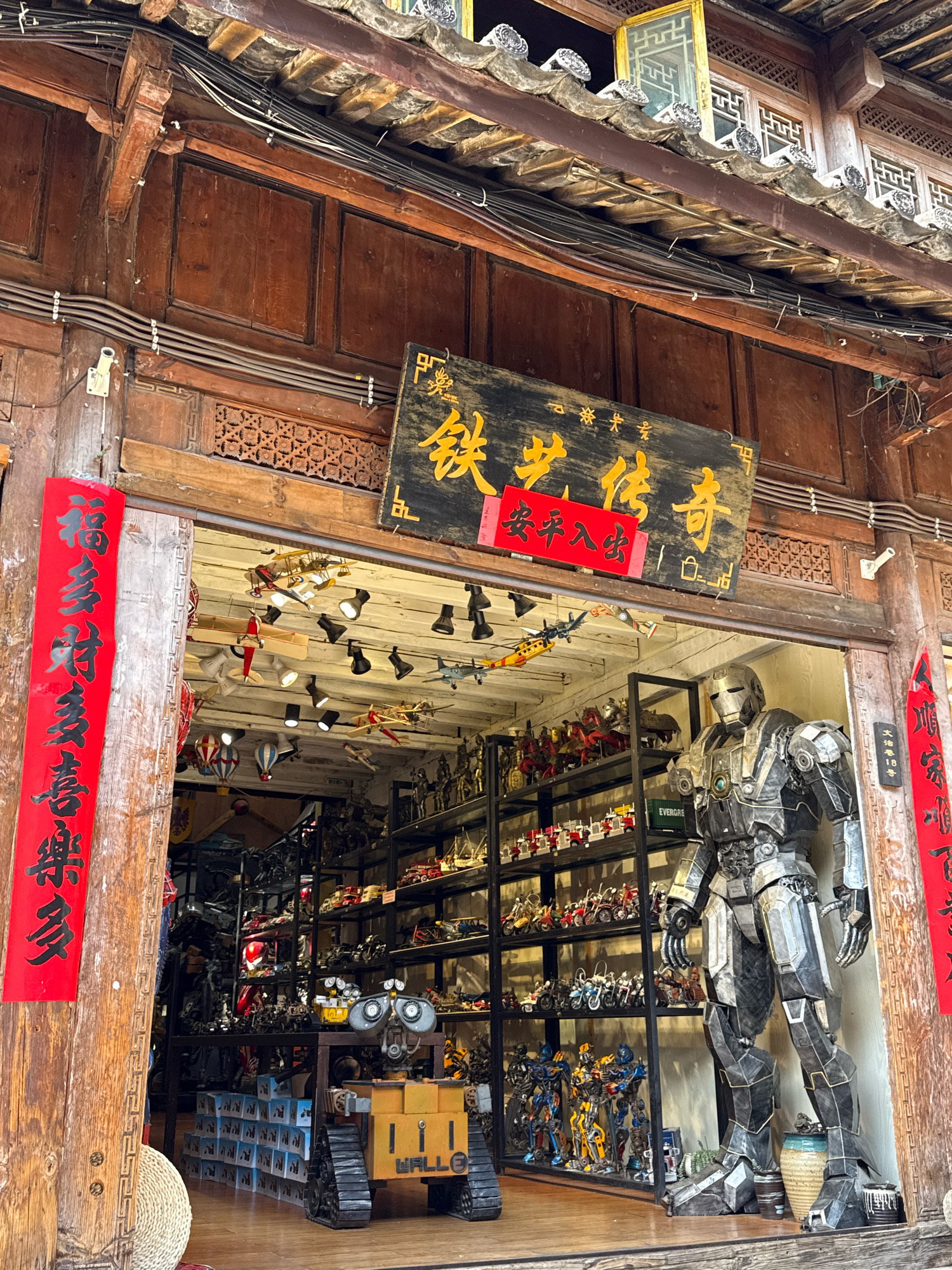 The image captures the storefront of a shop in Gucheng District, China, displaying a large collection of model vehicles and robots. The entrance is framed by traditional wooden architecture with intricate carvings and a tiled roof. A red banner with black Chinese calligraphy hangs vertically on the left side of the entrance, while a horizontal wooden sign with golden Chinese characters and a smaller red sign beneath it is mounted above the doorway.
Inside, the shop is densely packed with shelves filled with various miniature models, including airplanes, helicopters, motorcycles, and trucks. Prominently displayed in the foreground, to the right of the entrance, is a life-sized replica of a metallic robot, resembling a character from a science fiction film. To the left of the robot, a model of WALL-E, the animated robot character, is also visible, along with stacks of boxed merchandise. The interior lighting appears to be from spotlights suspended from the ceiling. The overall scene suggests a store specializing in collectible models and toys.