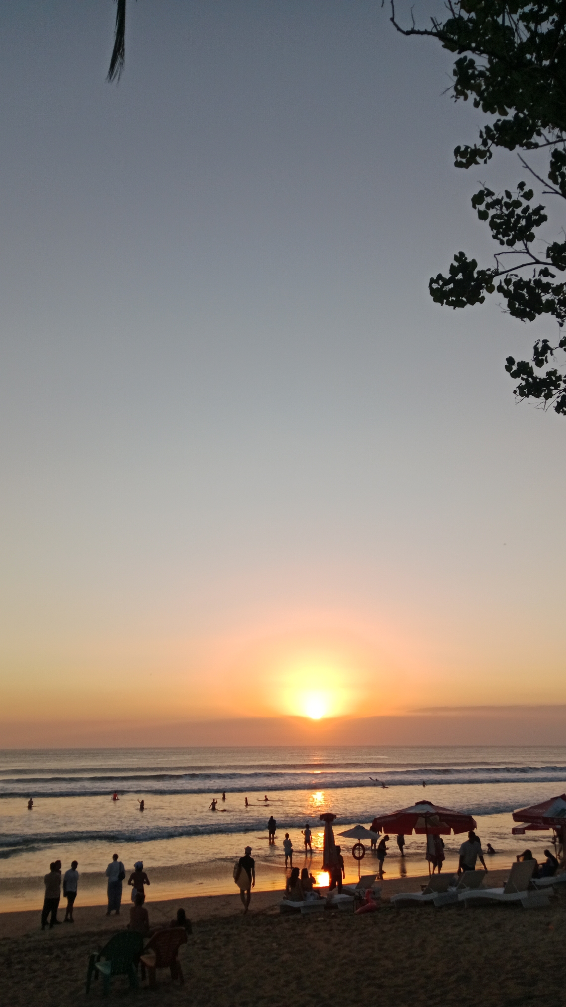 A sunset scene unfolds on Kuta Beach, Bali, Indonesia. The sun, a bright orb, dips towards the horizon, casting a warm, golden-orange glow across the sky and ocean. Gentle waves roll onto the sandy shore where numerous people are present. In the foreground, several individuals stand or sit on the sand, some with beach umbrellas providing shade over lounge chairs. Further back, more people are scattered along the waterline, with some wading in the shallow water or swimming. In the distance, the sun's reflection shimmers on the water's surface. A few trees with foliage are visible in the upper right and left corners of the image, framing the expansive sky.