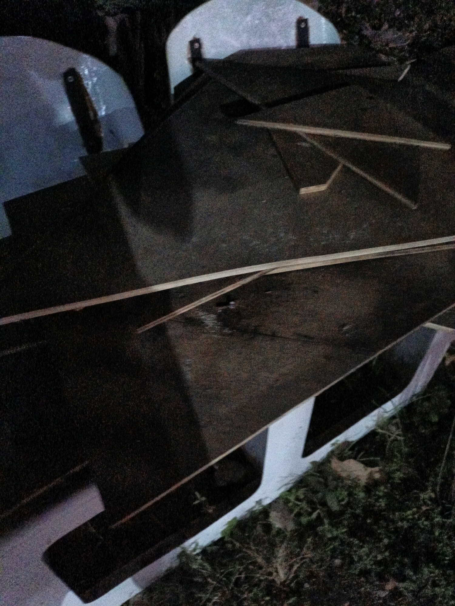 The image depicts an outdoor scene, possibly a backyard or a construction area, at night. Several large, dark sheets of material, likely plywood or particle board, are scattered on the ground and on top of what appears to be some sort of white, curved structures. These structures are partially visible and seem to be made of a smooth, solid material, possibly plastic or fiberglass. Thin wooden planks are also present, some laid across the larger sheets. The ground is covered with dry leaves and grass, indicating an outdoor environment. The overall lighting is dim, suggesting it is dark outside. There are no people or animals visible in the image. The location is Cikal, Indonesia.