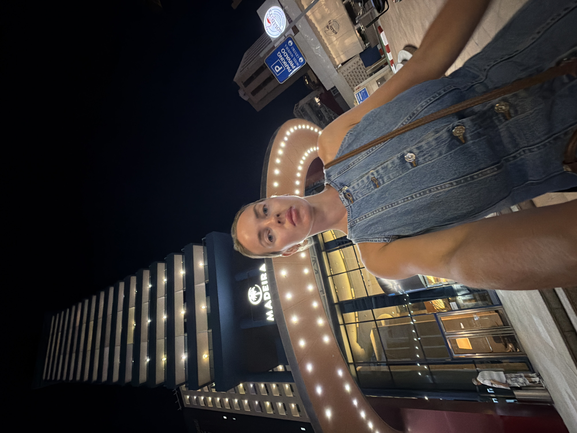 The image shows a woman taking a selfie at night in front of a hotel named "MADEIRA" in Benidorm, Spain. The hotel appears to be a tall, modern building with numerous lit windows, suggesting it is a significant structure in the city. The entrance to the hotel is grand, with a curved awning illuminated by a string of lights. To the right of the hotel, there are signs indicating parking, with one sign showing a blue "P" symbol and text in Spanish. Another sign displays the "HOLIDAYS" logo. The overall environment suggests an urban setting at night, with the bright lights of the hotel and surrounding buildings creating a lively atmosphere. The woman is wearing a denim dress and a bag, and her facial expression is neutral as she looks towards the camera. The scene is captured from a low angle, emphasizing the height of the hotel.