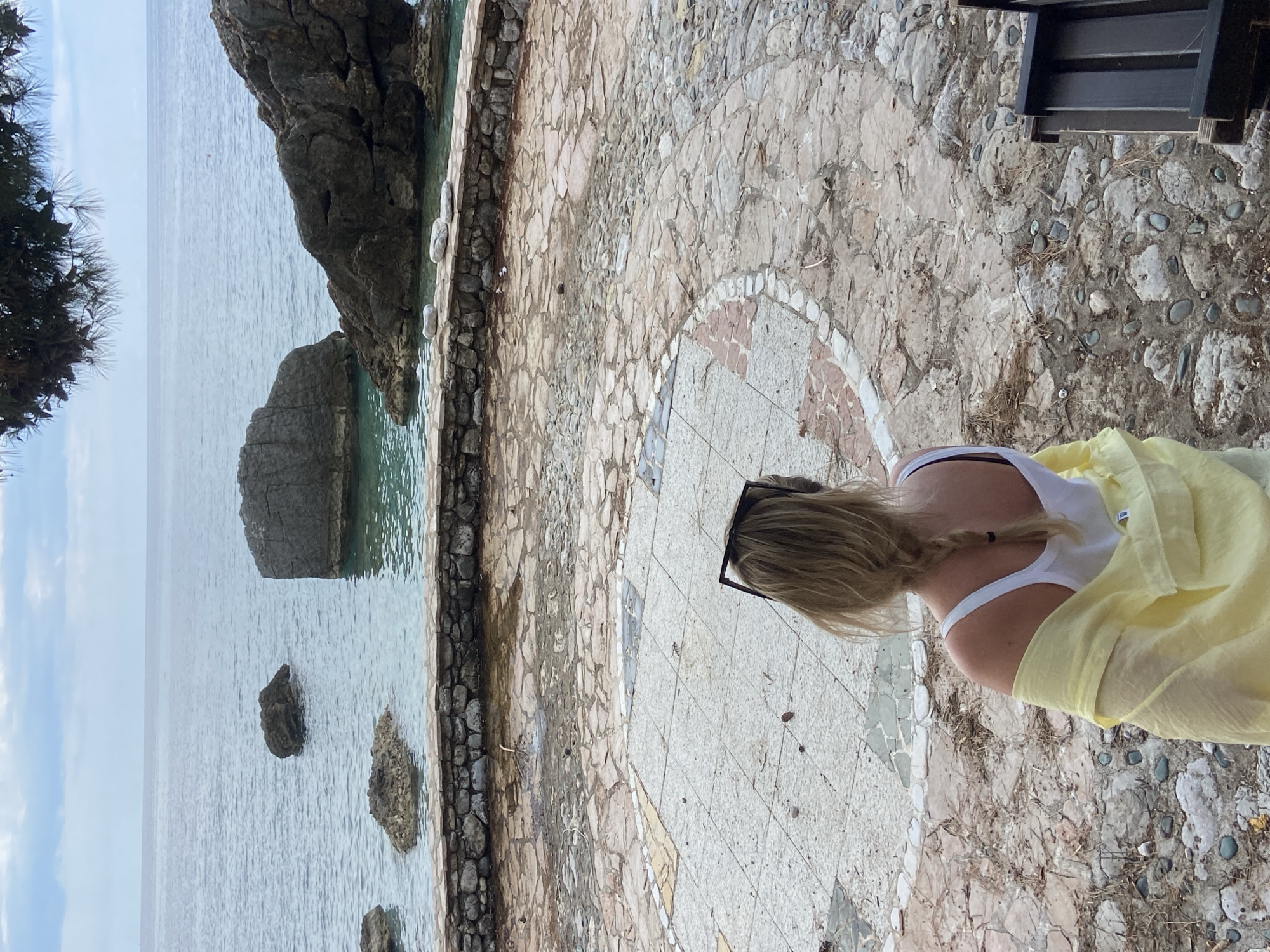 A woman is seen from a top-down perspective, standing on a stone-paved terrace overlooking the sea. The woman has blonde hair, a white tank top, and a light yellow shawl draped over her shoulders. Sunglasses rest on her head. The terrace, composed of various stones and tiles, features a circular pattern at the center. Beyond the terrace, the sea stretches out with rock formations emerging from the water. The location appears to be a scenic overlook, possibly in the city of Prijevor, Montenegro, given the presence of a rocky coast and seaside setting. A portion of dark-colored furniture is visible in the upper right corner of the image.