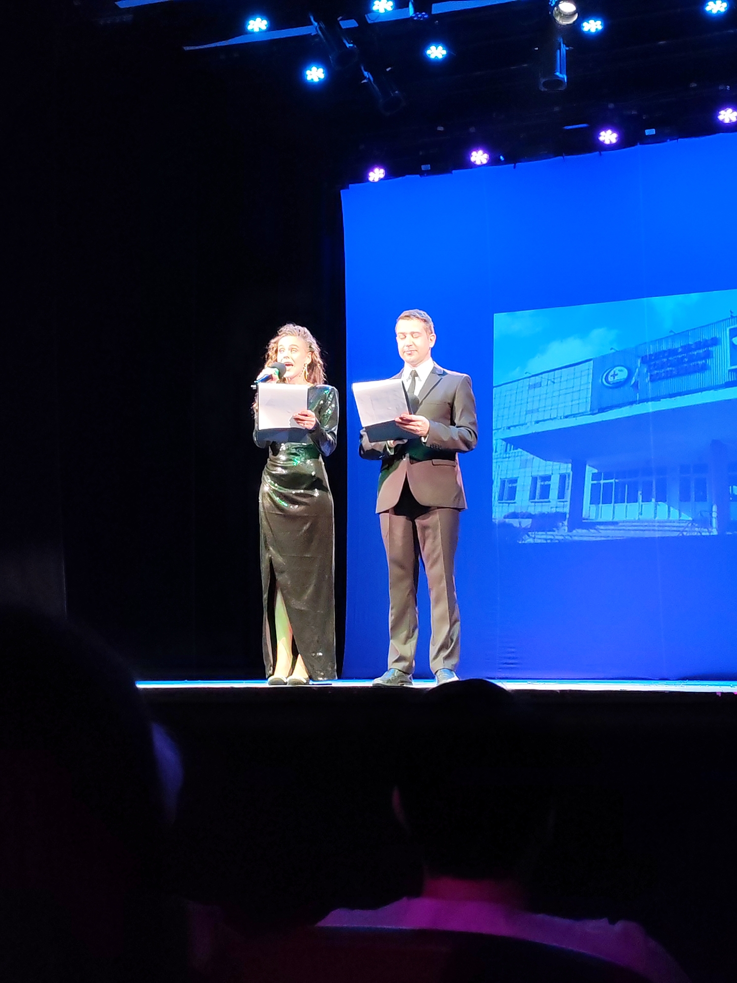 Two presenters, a woman in a sparkly black dress and a man in a dark suit, stand on a stage in front of a large blue screen in Chaikovsky, Russia. The woman is on the left, holding a microphone and a piece of paper. She has curly brown hair and is wearing a long, sparkly dress with a slit. The man is on the right, also holding a piece of paper. He has short brown hair and is wearing a suit and tie. The blue screen behind them displays an image of a building with a sky in the background. The stage is lit with blue spotlights, and the overall scene suggests a formal event.
