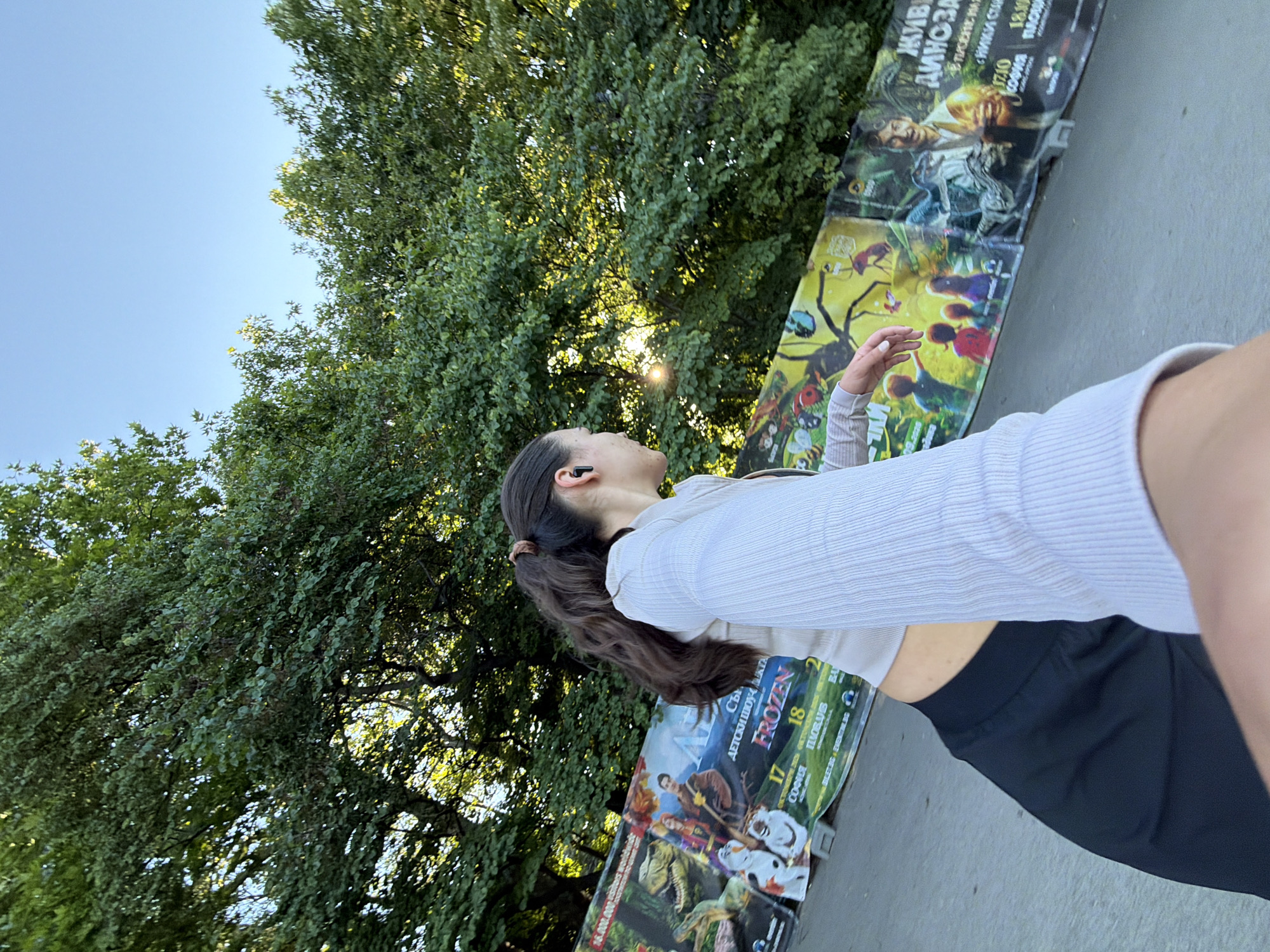 A young woman with dark hair in a ponytail and wearing black earbuds, a light-colored ribbed crop top, and black pants is standing outdoors in front of a large banner that appears to be advertising events. The banner displays colorful graphics, including dinosaurs, animated characters, and what looks like a movie poster for "Frozen." Text on the banner is partially visible and includes dates and locations, with "SOFIA" and "VARNA" being legible. Trees with green foliage form the background, and the sky is clear and blue. The sun is shining, creating lens flare effects. The overall impression is a casual outdoor setting, possibly a park or promenade, with promotional material displayed.