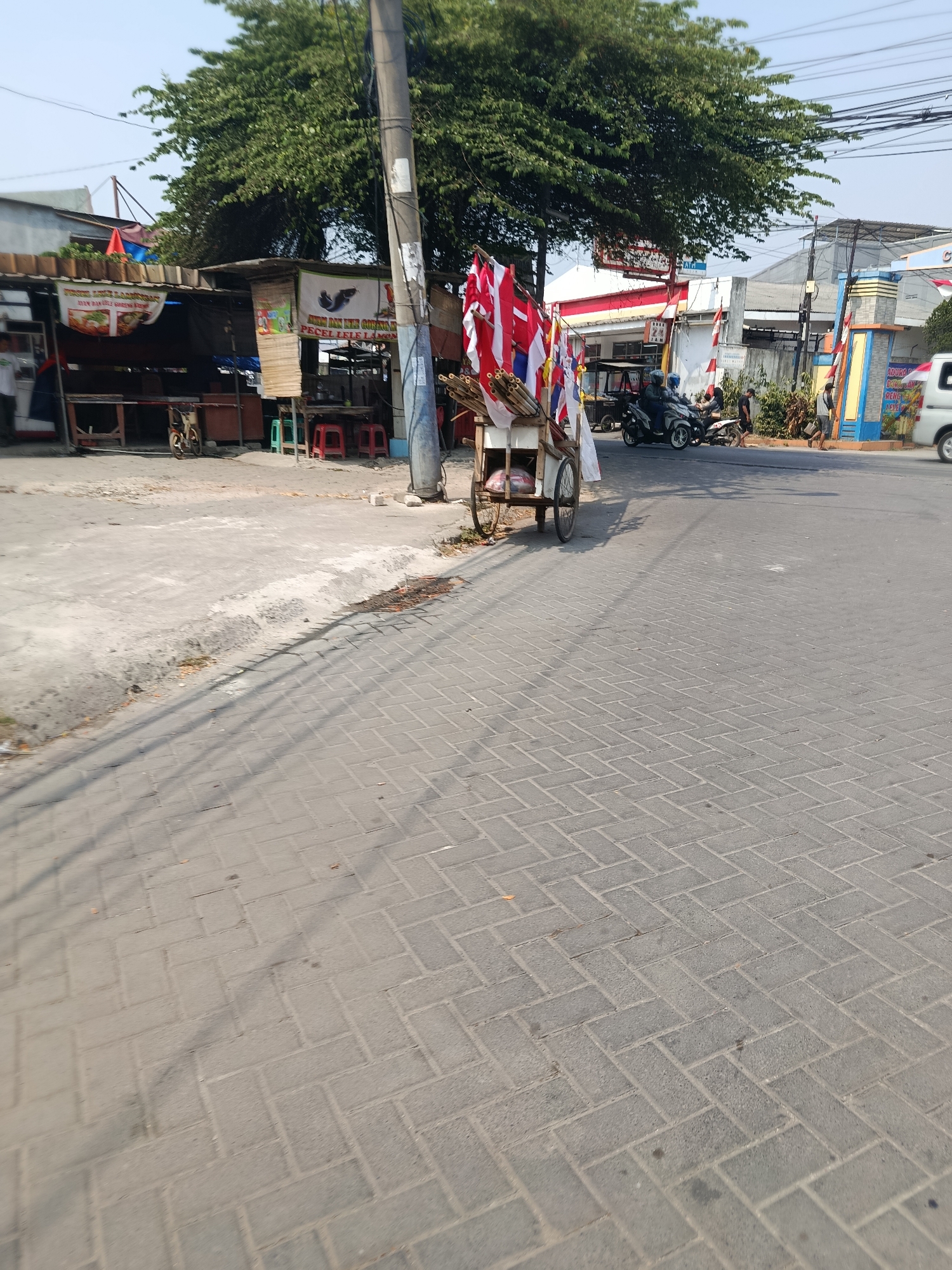 The image depicts a street scene in an urban environment, likely in Southeast Asia, given the presence of street food stalls and motorcycles. In the foreground, a paved road with interlocking brick patterns is visible, with a slight incline towards the right. A street vendor's cart, laden with what appear to be Indonesian flags and some sort of merchandise wrapped in plastic, stands on the left side of the road. The cart has two wheels and a handle for manual propulsion.
Behind the cart, a row of small, open-air food stalls lines the street. These stalls have simple structures with tarpaulin roofs and wooden tables and chairs, suggesting a local eatery. Signs in Indonesian are visible, advertising food items like "Pecel Lele" (fried catfish). A bicycle is parked near one of the stalls.
Further down the street, several motorcycles are parked and in motion. Two individuals are riding a motorcycle, and another motorcycle is stationary. People are walking around the stalls and the roadside. The background features more buildings, including what looks like a commercial establishment with a bright blue and yellow facade. Power lines crisscross the sky, and a large, leafy tree provides shade. The overall atmosphere suggests a bustling, everyday street scene in a city.