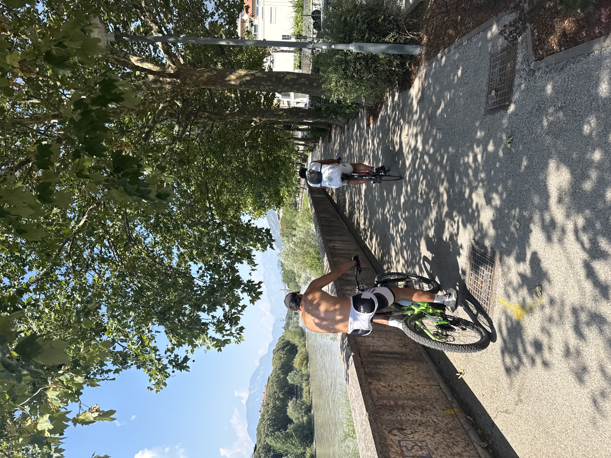 The image depicts a sunny day scene along the Adige River in Trento, Italy. Two individuals are riding bicycles on a paved pathway bordered by a stone wall and mature trees. The person in the foreground, seen from the rear, is a man with a bare torso, wearing white cycling shorts with black accents and a black baseball cap. He is riding a mountain bike with a neon green frame and black wheels. In the background, another cyclist, also seen from the rear, is wearing a white t-shirt, white shorts, and a black backpack. They are riding a road bike. The river flows to the left of the pathway, with a tree-lined bank and mountains visible in the distance. To the right of the pathway are more trees, a streetlamp, and buildings. The ground is paved and dappled with sunlight and shadows from the overhead trees. The overall impression is one of a recreational activity in an urban park setting.