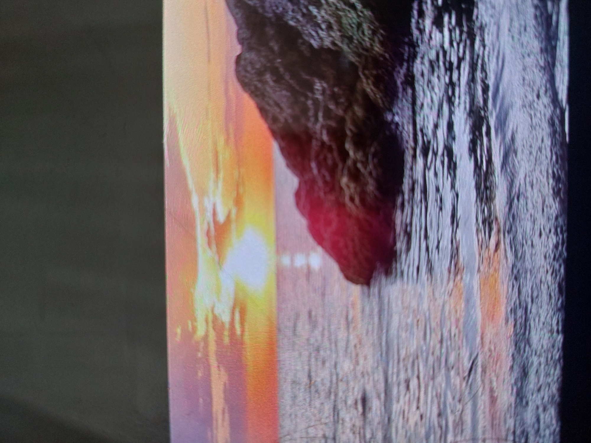 The image appears to be a close-up of a photograph or image on a screen, depicting a coastal scene at sunset. A rock formation is visible, with a textured surface and dark coloration. The sunset casts a warm, orange glow on the scene, with reflections appearing on the water. The water has a textured appearance, possibly indicating ripples or waves. The location is possibly Tsarevo, Bulgaria, based on the coastal nature of the scene.