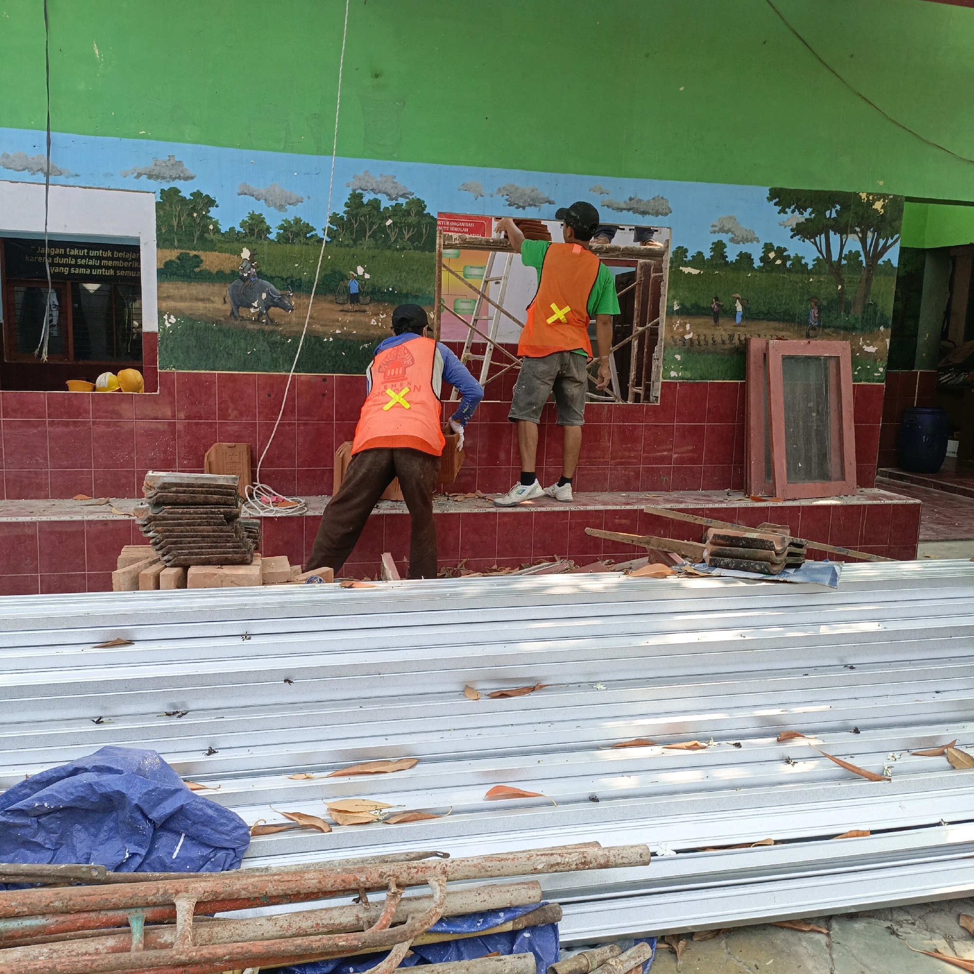 The image shows a section of a building under renovation, likely a school or community center, given the mural on the wall. Two construction workers, wearing orange safety vests, are visible. One worker is on the left, holding a brick and facing away from the camera, while the other worker is on the right, standing on a ladder and seemingly interacting with the wall, also facing away. The building has a green upper wall and a lower wall covered in red tiles. A mural depicting a rural landscape with trees, clouds, a water buffalo, and people engaged in farm work adorns the wall. Debris, including bricks, tiles, and metal framing, are scattered around the work area. Two yellow hard hats are placed on a ledge near a window. In the foreground, stacks of metal framing and a blue tarp suggest ongoing construction or demolition. The scene is outdoors, and the overall impression is one of a work in progress. The presence of the Indonesian language on signs and the style of the mural suggest the location is in Indonesia, specifically Salatiga, based on the provided context.