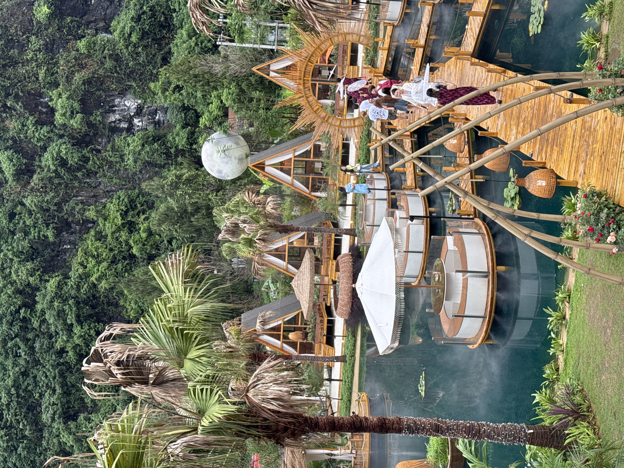 This image depicts a scenic view of a resort or park in Guilin, China, characterized by a blend of natural landscapes and crafted architectural elements. Lush green vegetation covers the backdrop, possibly a hillside or cliff, adjacent to a water feature (likely a pond or lake). Several uniquely designed structures are dispersed throughout the scene, including A-frame buildings with thatched roofs and a large spherical object, resembling a moon, positioned near one of the structures.
A wooden pathway or bridge winds over the water, connecting different sections of the resort. Several individuals are present on the pathway, appearing to be tourists or visitors, some with umbrellas, enjoying the setting. Additional elements include bamboo structures, decorative lighting, and meticulously landscaped gardens, indicating a well-maintained and aesthetically pleasing environment. Overall, the image showcases a blend of nature and designed architecture in the picturesque landscape typical of Guilin, China.