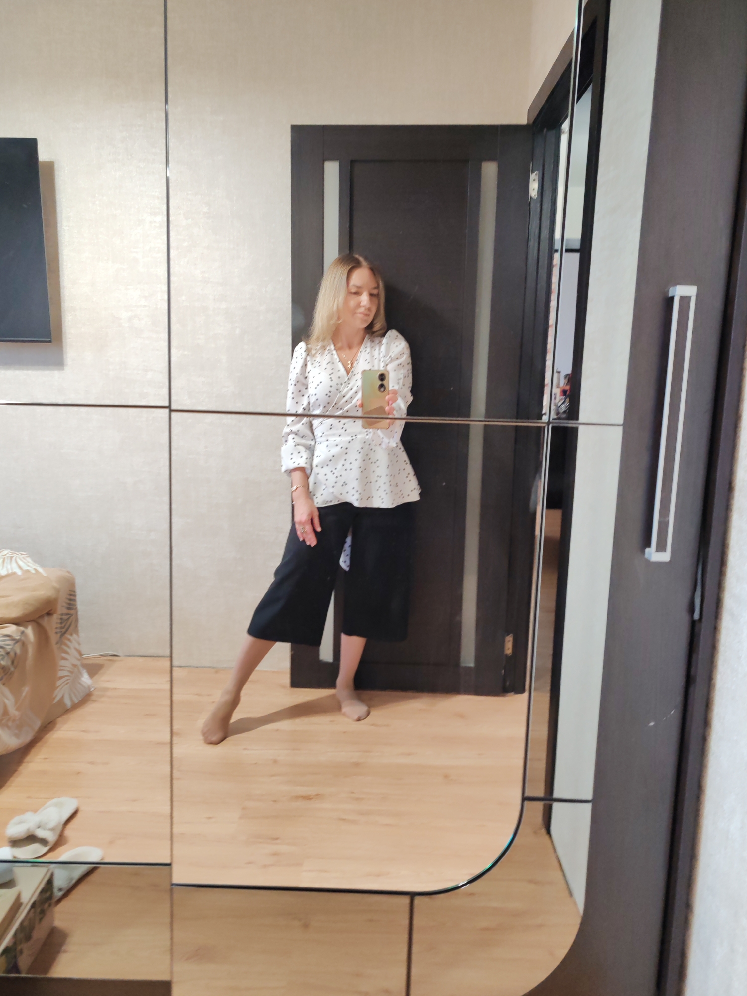 A woman is captured taking a mirror selfie indoors, likely in Petropavlovsk-Kamchatsky, Russia, during the daytime. She stands facing a large, multi-paneled mirror, which reflects her full-length figure. She wears a white V-neck blouse adorned with small black polka dots and black wide-leg culottes. Her blonde hair is parted on the side and falls past her shoulders. She holds a light-colored smartphone (possibly green or beige case) in her right hand, positioned to take the picture.
Her expression is relaxed, with a soft smile. She is barefoot, with her right foot (viewer's left) partially lifted, showcasing her light-toned skin. She wears a necklace with a cross pendant, a bracelet on her left wrist, and rings on her fingers.
The mirror's reflection reveals the interior of the room. Behind the woman, a dark brown door is visible. To the left of the reflection, a light-colored wall features a mounted black rectangular object, which appears to be a television. In the lower left, part of a bed or sofa with a light-colored, patterned throw is reflected. On the light wood-patterned floor, a pair of white, fuzzy slippers are also visible in the reflection. The mirror itself is divided into several rectangular sections by thin vertical and horizontal lines. There is no visible text in the image.
