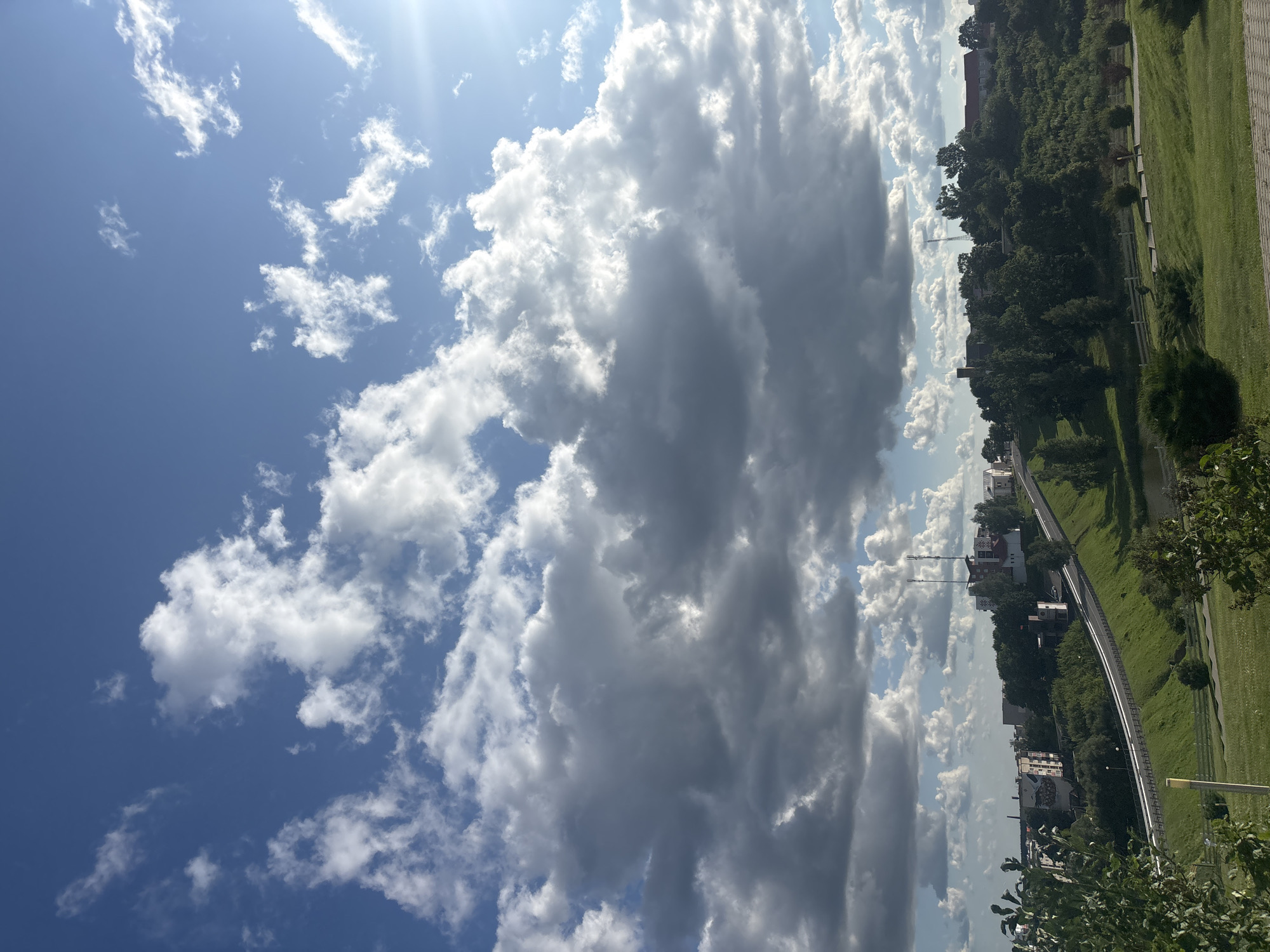 The image depicts a landscape view of the city of Kapyĺ, Belarus, under a dynamic sky. The foreground features a well-maintained grassy area with a path and some bushes. A sloping, green hillside dominates the midground, with a paved road winding its way up and across it. Along the top of the hill, buildings and structures, including houses and what appear to be communication towers, are visible amongst dense green trees. The background is filled with a vast blue sky characterized by a mix of dramatic, dark grey storm clouds and bright white cumulus clouds. The sun is visible on the right side, casting light and creating a lens flare. The scene captures a transitional moment, possibly before or after a rainfall. There are no people or animals visible in the frame.