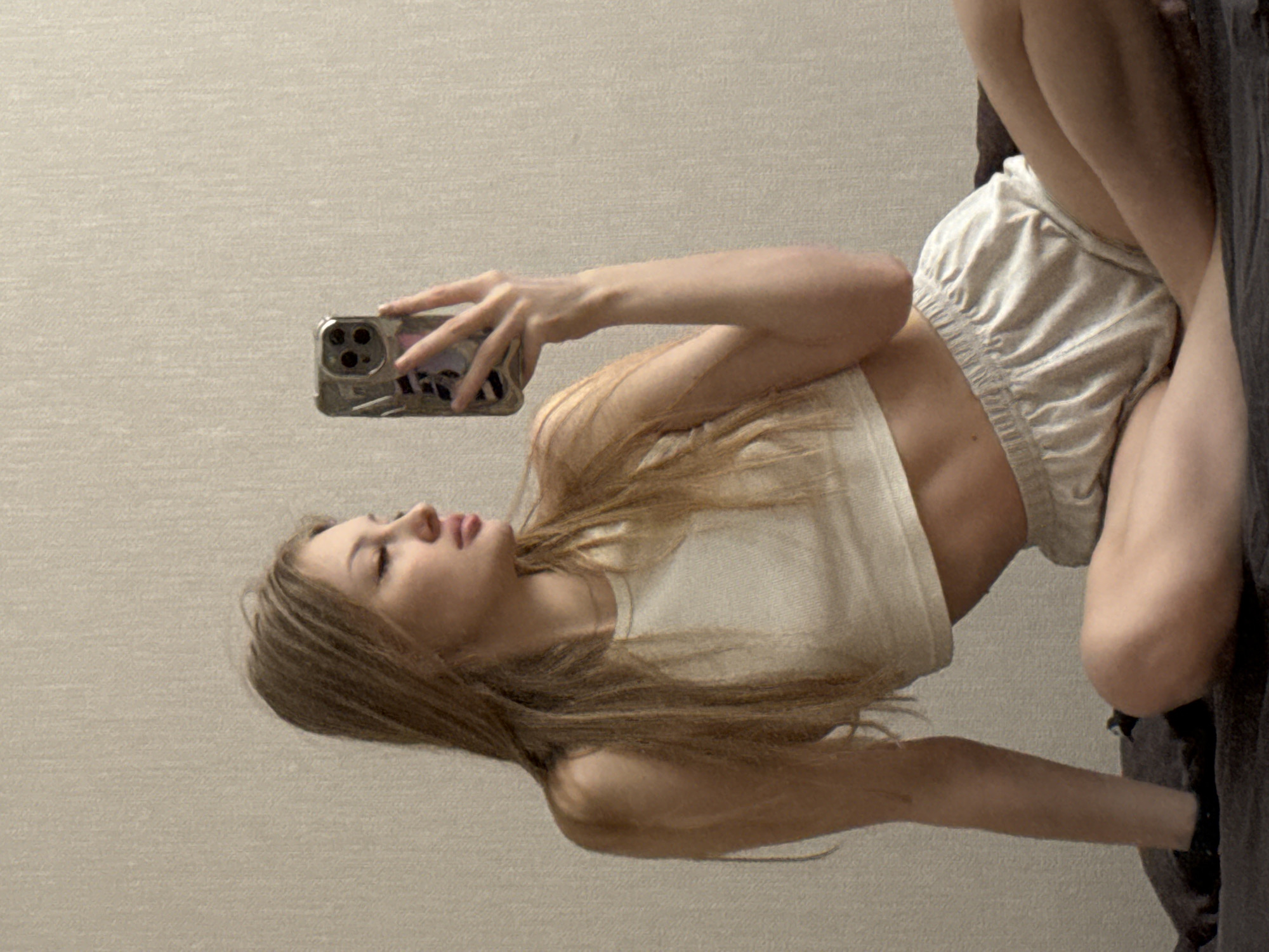 A young woman is the central subject of this image, captured in what appears to be a self-portrait or mirror selfie taken with a smartphone. The image is oriented sideways, with the woman appearing to lie on her left side or recline at an angle. She holds a smartphone, likely an iPhone given the distinctive triple camera module, in her right hand, directing her gaze towards its screen.
She has long, light brown or blonde hair, cascading down. She is dressed in a light-colored, possibly white or cream, crop top that exposes her midriff, and matching light-colored shorts with an elasticated waistband. Her body posture suggests a relaxed pose, with her legs bent.
The background is a plain, light beige wall with a subtle textured pattern. A darker, soft surface, possibly a bed or couch, is visible in the lower right portion of the frame, where she appears to be situated.
Her facial expression is neutral, with her lips slightly parted. The phone case she holds is reflective, obscuring any clear details or text that might be present on it. No discernible text is visible elsewhere in the image. The setting is indoors, with no specific time of day or weather indicated. The location context provided is Perm, Russia.