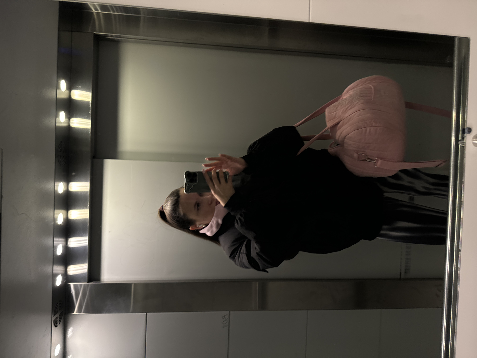 A woman is taking a selfie in a mirror. She is wearing a black jacket and has a pink backpack over her shoulder. The mirror is in a public space, potentially an elevator, with visible lights. A partial inscription is visible on the mirrored wall: "B09" with other illegible scribbles. The location is Odintsovo, Russia.