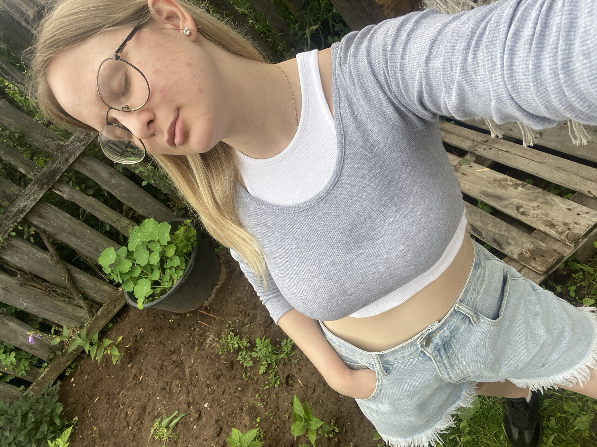 A young woman with blonde hair and glasses is standing in a garden, with a wooden fence in the background. She is wearing a grey long-sleeved crop top, a white tank top, and light-wash denim shorts with a frayed hem. Her right hand is in her pocket. The ground is covered in dirt and some greenery, with a black pot containing plants and a wooden pallet visible. The location appears to be Kauchuk Stantsiya, Russia.
