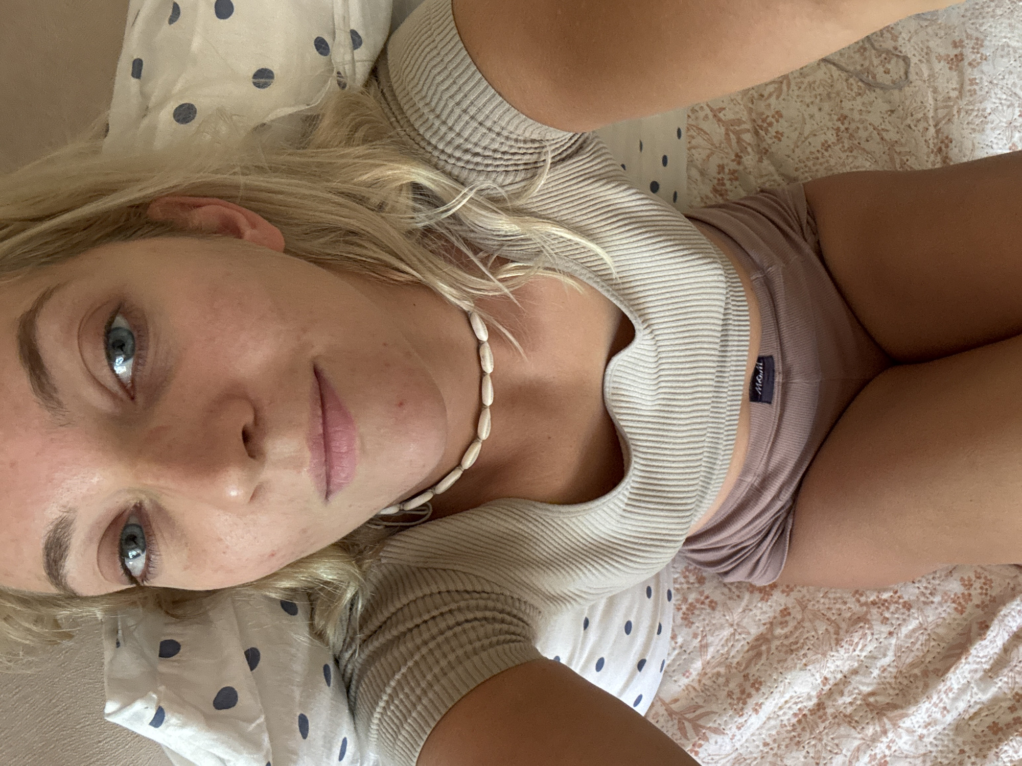 A blonde woman lies in bed. She is wearing a light-colored ribbed shirt and shorts. She has blue eyes and is wearing a necklace. The bed is covered in patterned sheets. There is no evidence to suggest this photo was taken in Minsk, Belarus.