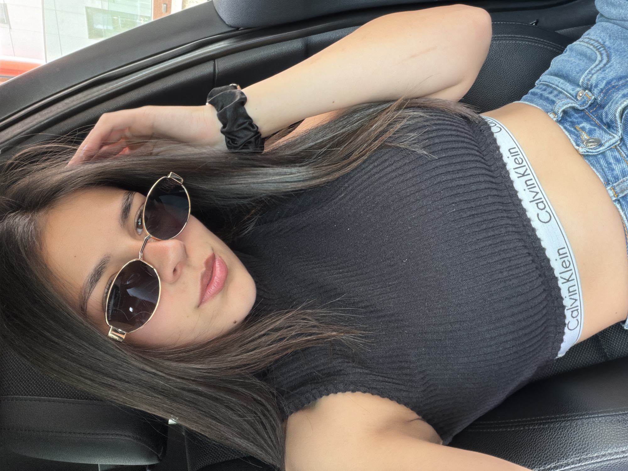 A young woman is lying down, possibly in the reclined seat of a car, and looking towards the viewer. She has long, dark brown hair, with some strands draped over her shoulder and arm. Her face is partially covered by gold-framed oval sunglasses, and her lips are slightly parted in a relaxed expression.
She is wearing a black ribbed crop top, which reveals a white elastic waistband underneath. The waistband clearly displays the text "Calvin Klein" repeated in black capital letters. Her lower body, visible on the right side of the frame, is covered by light-wash denim jeans or shorts. A black scrunchie is worn on her left wrist. The interior of the vehicle features black leather seats and a dark headliner, indicating an indoor setting within the car. The lighting suggests it is daytime. This scene is located in Gabrovo, Bulgaria.