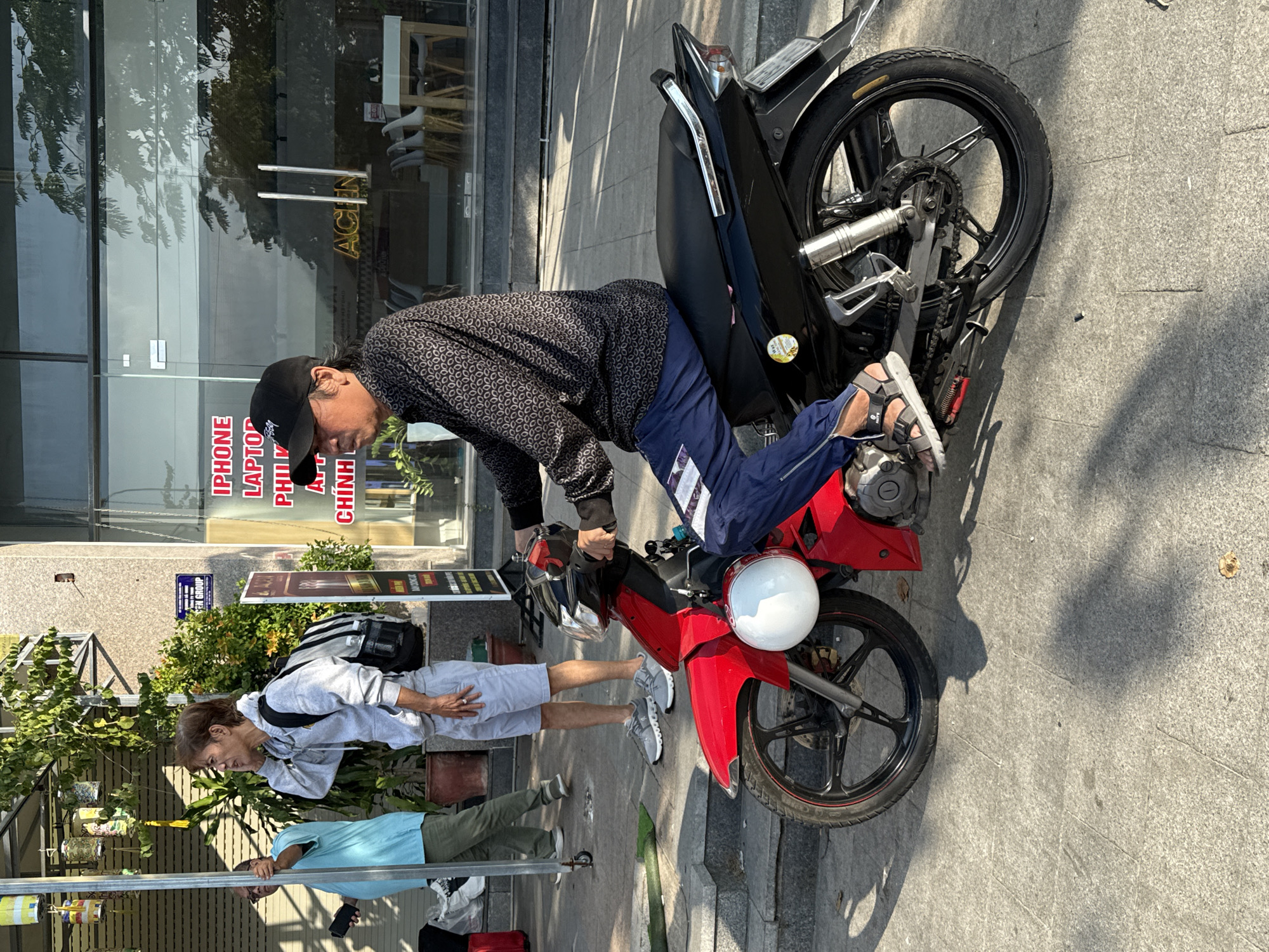 The image shows a street scene in Nam Nha Trang Ward, Vietnam. A man wearing a black baseball cap, a patterned long-sleeved shirt, and blue pants is riding a red and black motorcycle. He is wearing sandals. A white helmet is attached to the front of the motorcycle. In the background, another person in a light blue shirt and olive pants is standing and looking to the side, holding a black object. A third person in a grey hoodie and light grey shorts is standing next to the person in the blue shirt. Behind them is a building with a glass facade and signage. The ground is paved with stone tiles, and there is a shadow of the motorcycle and riders on the ground.