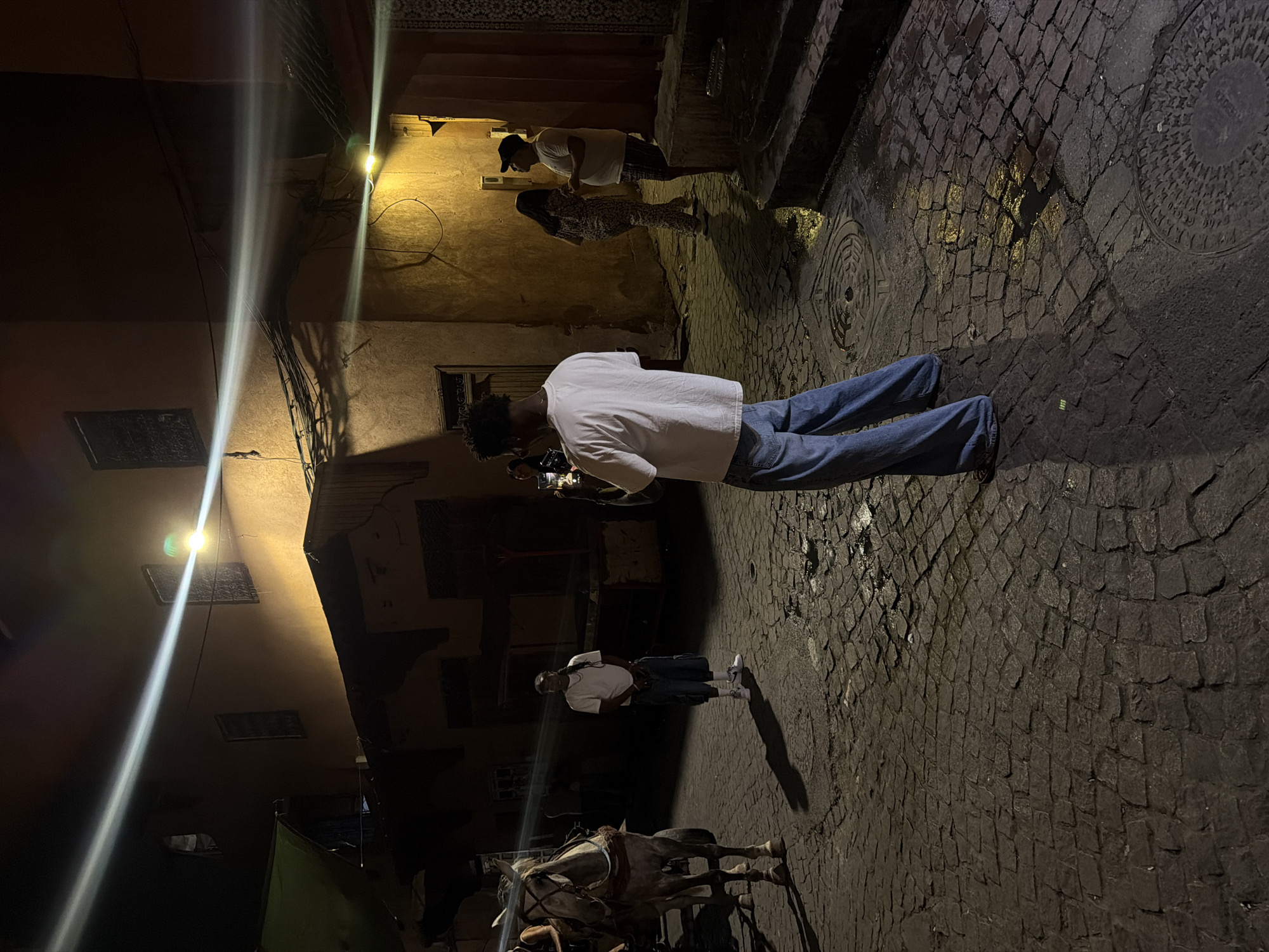 In Marrakesh, Morocco, at night, a street scene unfolds with cobblestone paving reflecting artificial light. The warm glow from buildings illuminates the narrow alley, highlighting architectural details and the presence of overhead wiring. In the foreground, a person stands with their back to the camera, dressed in a white t-shirt and loose-fitting blue jeans, holding a smartphone. To their left, a white horse, harnessed to a carriage, stands still. Further down the street, other individuals are visible. One person, wearing a white t-shirt and shorts, stands near a building, while two other figures, a woman in a patterned dress and a man in a t-shirt and cap, converse. The overall atmosphere is dimly lit, with strong directional light creating dramatic shadows.