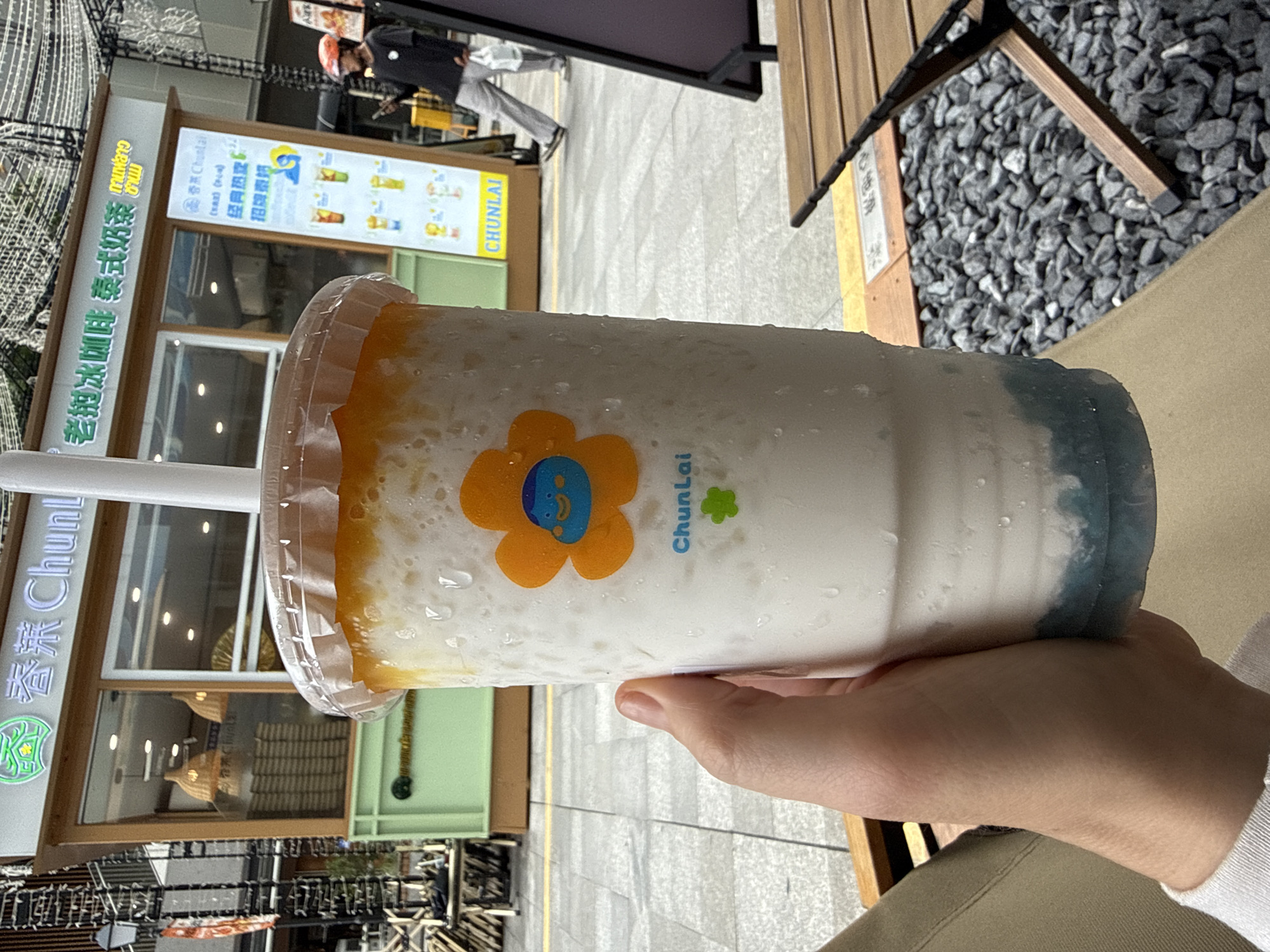 The image shows a close-up of a person's hand holding a large, clear plastic cup filled with a layered beverage. The drink appears to have a white, milky base with a yellow-orange layer at the top and a blue layer at the bottom. The cup has condensation on the outside and features an orange flower sticker with a blue cartoon face. A white straw is visible extending from the top of the cup.
In the background, a storefront with the words "春茉 ChunLai" is visible, along with Chinese characters. The storefront has a green base and a wooden frame. A sign with drinks pictured and the text "CHUNLAI" is also present. A person wearing a black long-sleeved shirt, grey pants, and an orange helmet is walking away from the storefront. The scene appears to be an outdoor seating area with cobblestone ground and wooden furniture.
The text on the storefront and sign indicates this is likely a "ChunLai" establishment, a brand known for beverages, possibly in Huizhou, China, given the use of Chinese characters and the context of Asian street scenes often associated with such brands. The overall environment suggests a commercial area, perhaps a street or plaza with shops.