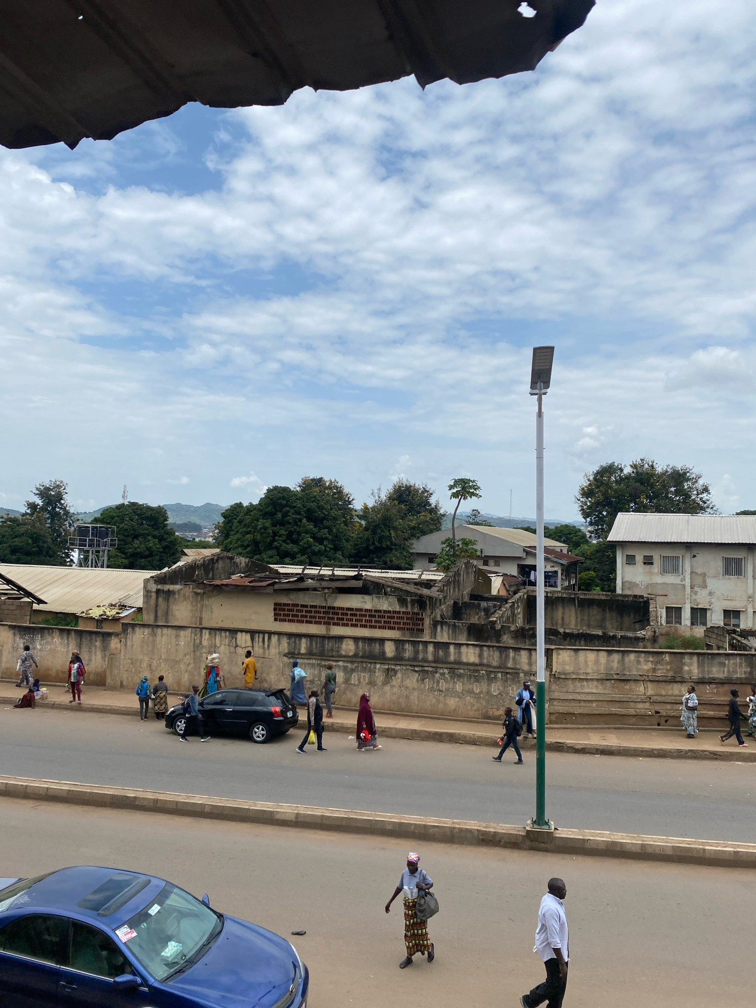The image depicts a street scene in Jos, Nigeria. A light pole with solar panels stands prominently in the middle of the frame. People are walking on the sidewalk and crossing the street. A black car is parked on the side of the road. In the foreground, a blue car is parked. Buildings and trees are visible in the background under a cloudy sky.