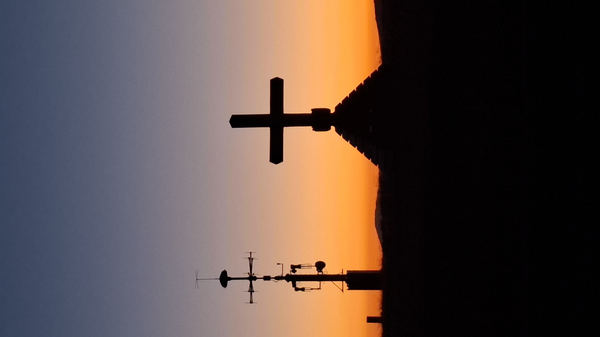 This is a vertical, eye-level, full-frame shot of a silhouette of two crosses against a gradient sunset sky. On the left is a tall, multi-layered structure with what appears to be an antenna or weather monitoring equipment. On the right is a large, prominent cross silhouetted on top of a tiered structure. Both objects are set against a horizon line of low hills. The sky transitions from a deep blue at the top to a bright orange near the horizon. The image was taken from the vicinity of Waldbach-Mönichwald, Austria. There are no people or animals visible in the image.