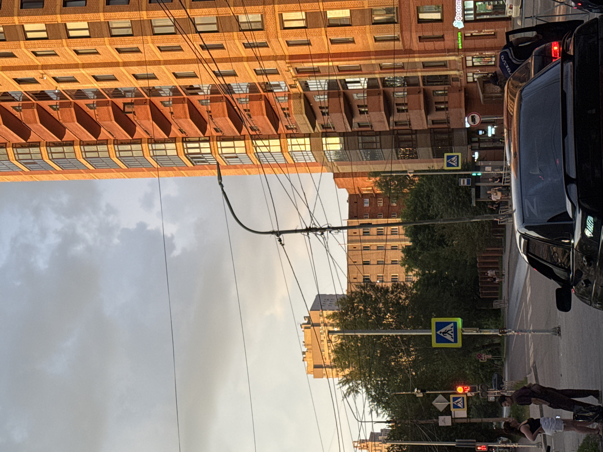This image captures an urban scene at street level, likely in Novosibirsk, Russia, indicated by the presence of Cyrillic signage. The foreground is dominated by a black car, partially obscuring the view of a street intersection. A pedestrian crossing with a blue and yellow diamond-shaped sign depicting a person walking is visible. Traffic lights show a red light, and a yellow pedestrian crossing sign is also present.
Two individuals, a woman in a black tank top and white shorts, and a man in dark clothing, stand near the pedestrian crossing. Further in the background, a tall, modern, brick-faced building with numerous balconies and glass-fronted sections is prominent, bathed in warm, evening sunlight. Across the street, another multi-story building with a pale facade and many windows is visible. Trees line the street, some of which are dense with green foliage. Overhead, power lines crisscross the sky, which is a mix of grey clouds and a lighter, possibly dusk-lit sky. A street lamp is also visible on the right side of the frame. The overall impression is one of a typical city street during the late afternoon or early evening.