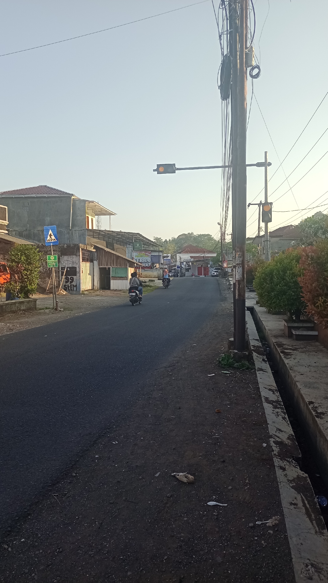 A roadside view in Karyamandala, Indonesia, captures a street scene at what appears to be dawn or dusk, with a clear sky and warm, low-angled sunlight. The paved road, slightly uneven, curves gently into the distance, flanked by a concrete drainage ditch on the right. To the left of the road, a few residential or commercial buildings are visible, some appearing to be under construction or renovation with exposed concrete and incomplete roofing. A blue pedestrian crossing sign is mounted on a pole near the left edge of the frame, accompanied by a green sign below it.
Several motorcycles are present. One person is riding a motorcycle down the road in the foreground, while another is further ahead, also on a motorcycle. In the background, more vehicles, including cars and motorcycles, are visible near buildings.
A prominent wooden utility pole stands on the right side of the road, heavily laden with wires and electrical equipment. Attached to this pole, and extending over the road, is a traffic light displaying a yellow signal. Another traffic light, also appearing yellow, is visible on a separate pole further down the road.
The ground beside the road is a mixture of dirt, gravel, and scattered debris, including what appears to be a dead bird and some small plastic items. Lush green and reddish-orange foliage can be seen in planters along the right side of the drainage ditch, adding a touch of color to the scene. The overall impression is of a typical semi-urban or rural Indonesian street.