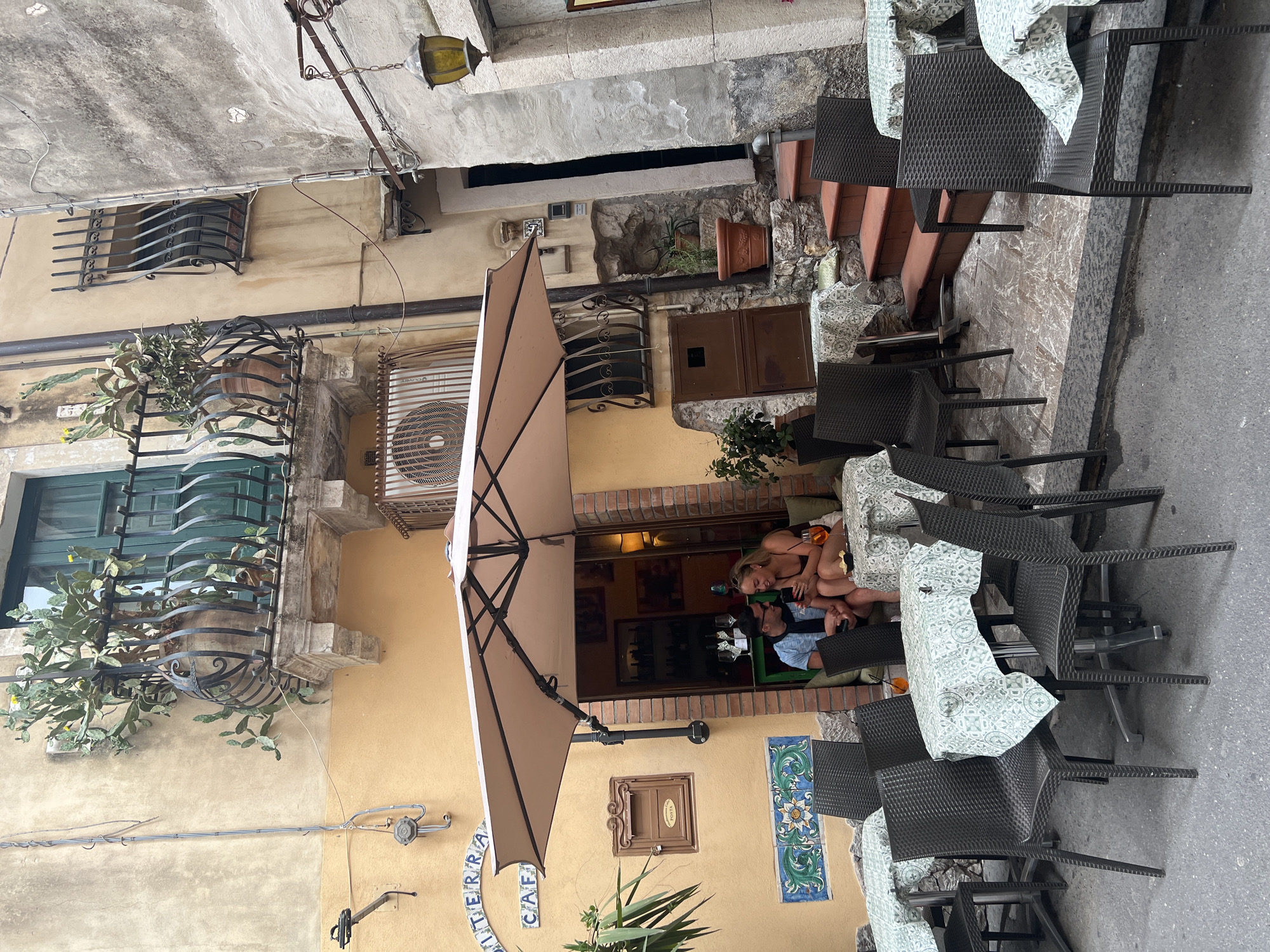 This is a medium shot of an outdoor cafe in Taormina, Italy. The cafe has tables and chairs set up on a cobblestone street. A couple is sitting at one of the tables, the woman is smiling at her phone while the man looks at it. The cafe has a large umbrella providing shade for the tables. The buildings surrounding the cafe are old and have a rustic appearance. There is a balcony with a potted plant on the building to the left of the cafe. The cafe sign reads "TERRA CAFE". The overall atmosphere is relaxed and inviting.