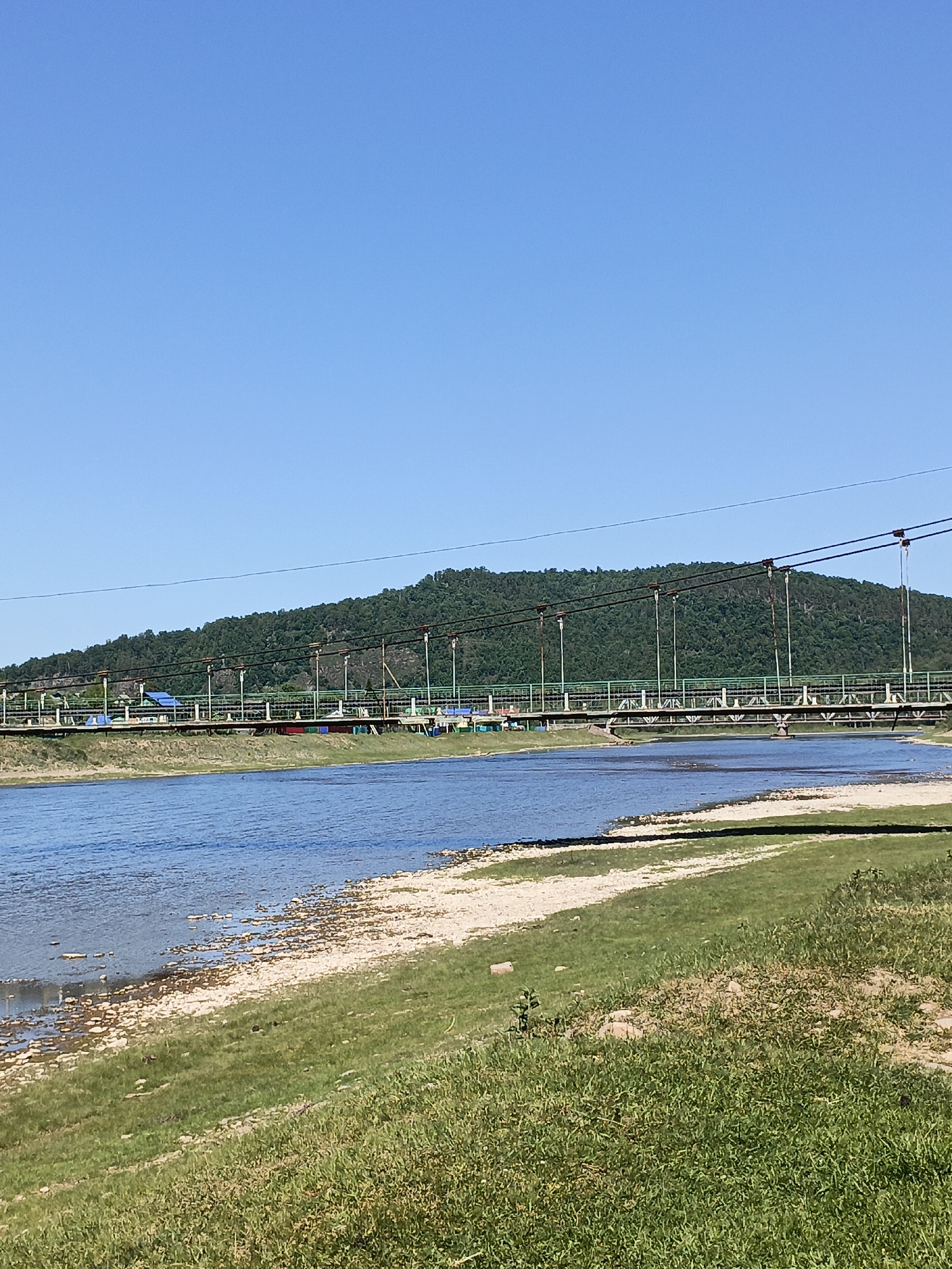 The image shows a low-lying pedestrian suspension bridge spanning a river in Gabdyukovo, Russia. The bridge has a green railing and lights above. The river is wide, with a shoreline of grass and sand on the near bank and a wooded hill in the background. The sky is clear and blue.