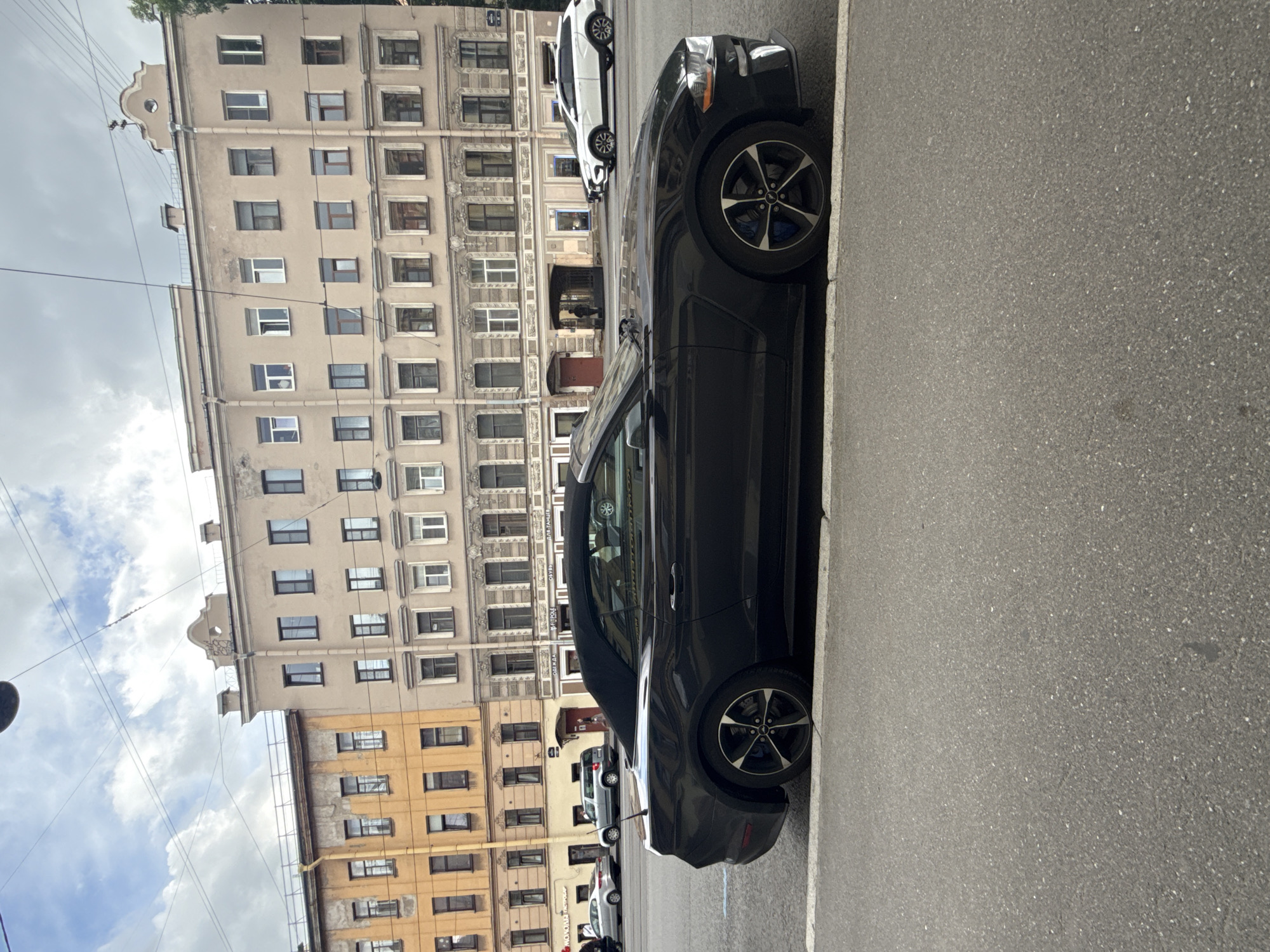 A black convertible Mustang is parked on a city street in Saint Petersburg, Russia, in front of a row of multi-story buildings. The buildings have a mix of beige and yellow facades with many windows. The sky is cloudy with visible power lines. Other cars are visible on the street, including a white SUV further down. The Mustang's black convertible top is down, and its wheels have black rims with blue brake calipers. The street is asphalt with a white line marking the parking area. The overall scene depicts a street view in an urban environment under an overcast sky.