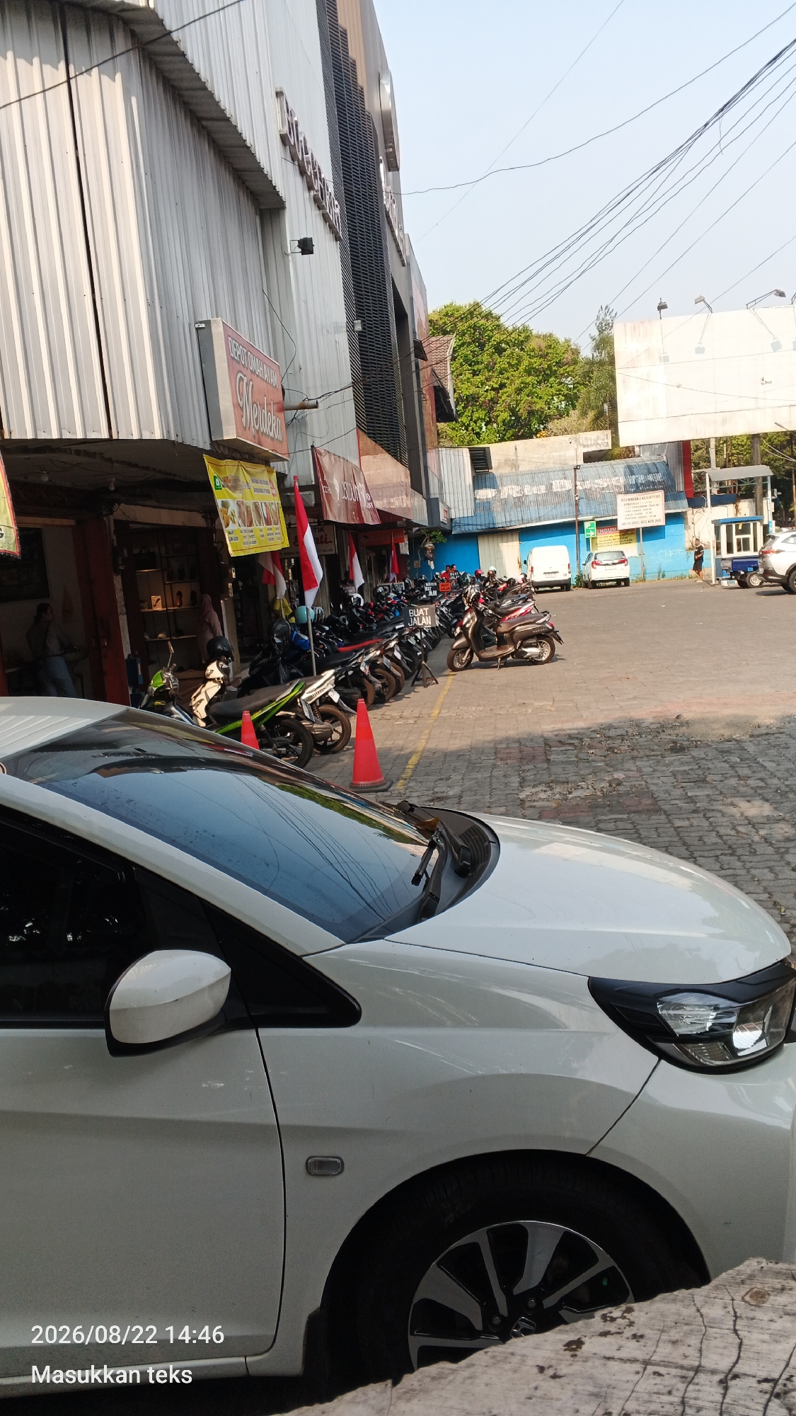 In Malang, Indonesia, on a sunny day, a white car is parked in the foreground, partially obscuring a row of parked motorcycles. The motorcycles are lined up in front of shops with corrugated metal facades and awnings. Some of the shops display red and white Indonesian flags. Beyond the shops, a street is visible with several other cars and some people. The background includes a large billboard, trees, and a clear sky. The time stamp on the image indicates August 22, 2026, at 2:46 PM.