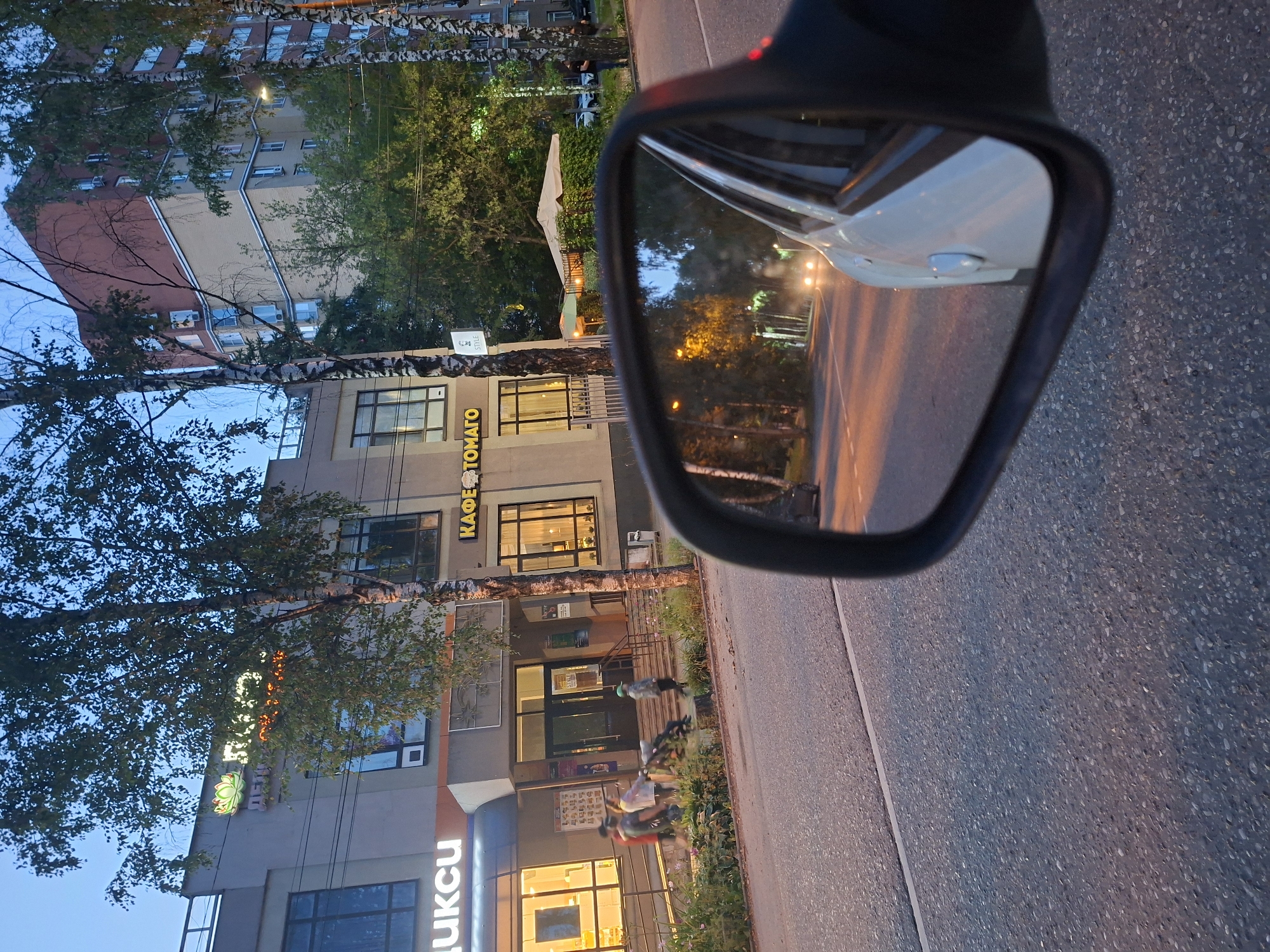 The image, taken in Aprelevka, Russia, captures a street scene with a focus on a building and its reflection in a car's side mirror. The building on the left features illuminated signage that reads "ЦИКСИ" and "КАФЕ ТОМАГО" (Cafe Tomato). Several people are visible walking along the sidewalk in front of the building. The reflection in the side mirror shows a road with approaching headlights, suggesting the photo was taken from a moving vehicle. Trees line the street, and a tall apartment building is visible in the background. The lighting indicates it is either dusk or early evening.