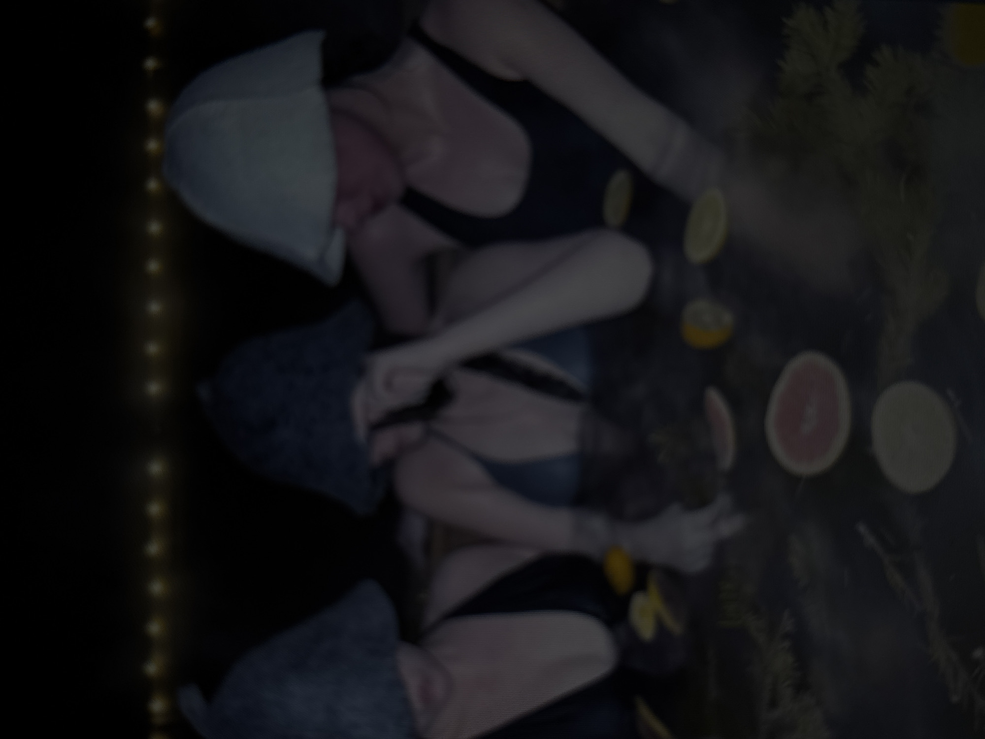 The media file shows three people submerged in water, possibly a hot tub or pool, with a dark background and some faint yellow string lights on the left side. The subjects are partially visible and appear to be in a relaxed state. One person is wearing a white cap, another a dark bathing suit and a gray cap, and the third is wearing a dark bathing suit and a gray cap. Sliced fruits, including what appear to be grapefruits and lemons, are floating in the water. There are also some leafy branches or herbs submerged. The overall impression is one of a spa-like or wellness setting. The lighting is dim, suggesting it might be nighttime or an indoor setting with low light. The water appears calm. No text is visible in the media file. Given the location context of Yerevan, Armenia, this could be a personal spa experience or a commercial establishment.
