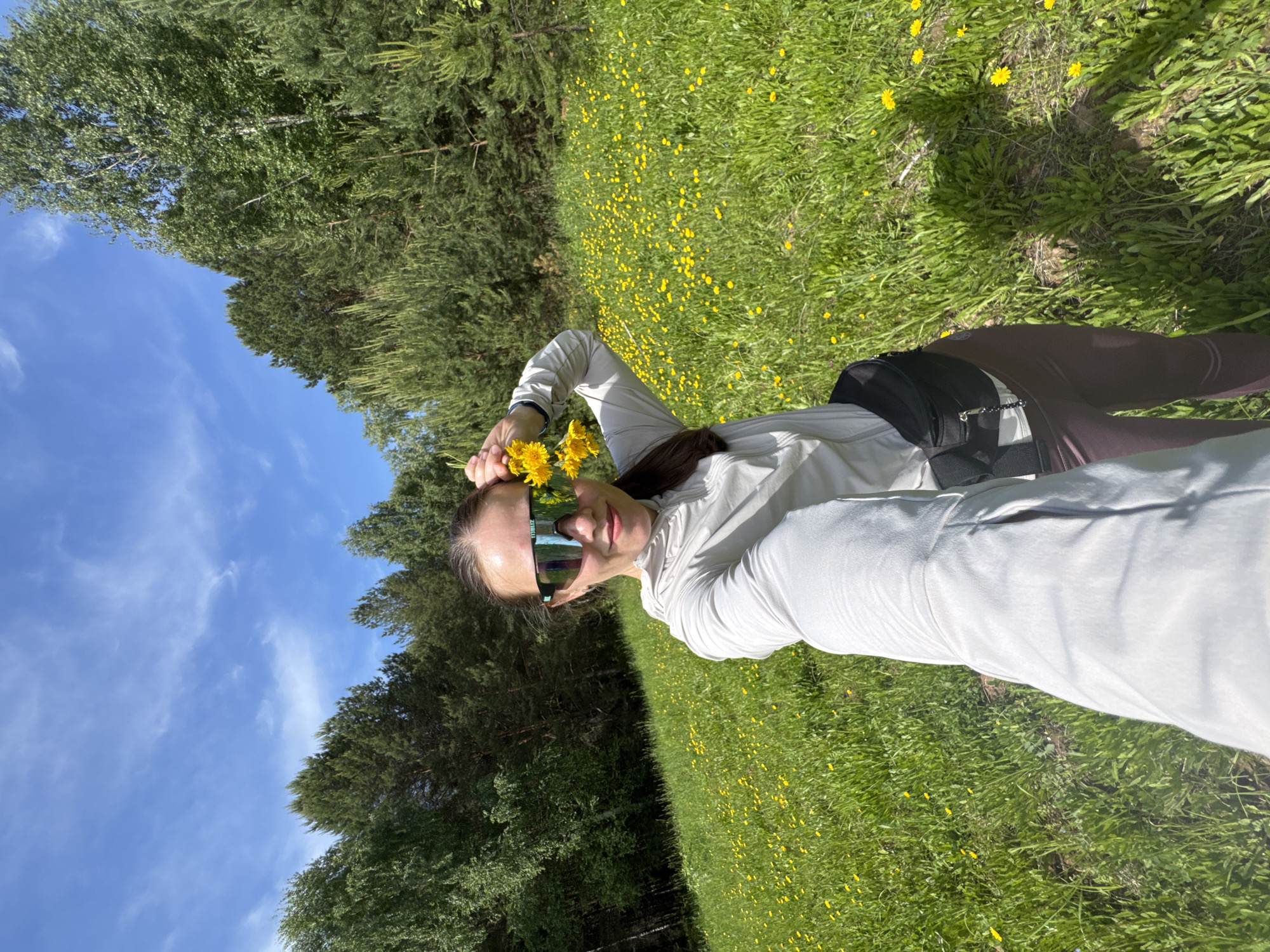 This is a vertically oriented outdoor selfie taken in bright daylight in Chaikovsky, Russia, depicting a person in a field of dandelions.
**Subjects:** The primary subject is a woman with long, dark hair, seen from the waist up. She is wearing light-colored, long-sleeved athletic wear, dark leggings, and large, reflective sunglasses. Her left hand is holding a small bouquet of freshly picked yellow dandelions near her head. The background features a vibrant green grassy field densely dotted with yellow dandelions, with a line of various green trees (including some with lighter bark like birch) forming a natural boundary behind her. Above the trees, a clear blue sky with sparse white clouds is visible.
**Activity or event taking place:** The woman appears to be posing for a selfie or self-portrait, looking directly at the camera with a slight smile. She is engaged in picking or holding dandelions, suggesting a leisurely outdoor activity in a natural setting.
**Notable details:** The time of day is clearly sunny, indicated by the bright illumination and visible shadows on the grass. The weather is clear and pleasant. The natural environment, with abundant dandelions and trees, suggests a spring or early summer scene. The woman's expression conveys a sense of calm and enjoyment. The sunglasses have the word "BLIZ" visible on the left temple. The image is taken with a tilted horizon, giving it a dynamic angle.