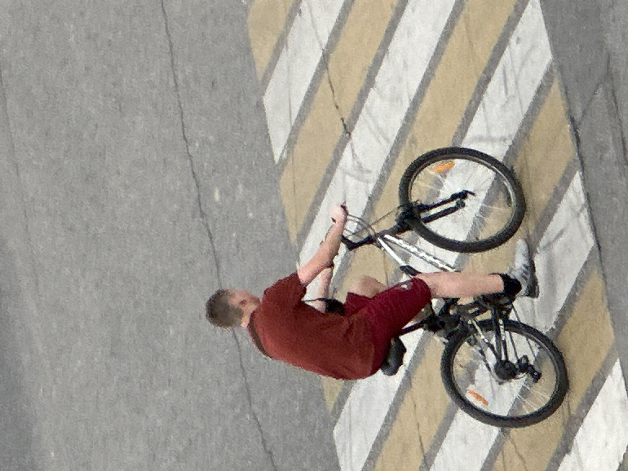 The image shows a person riding a bicycle across a crosswalk in Podolsk, Russia. The person is wearing a red t-shirt and red shorts. The bicycle is a mountain bike with black handlebars and a silver frame. The crosswalk is marked with white and yellow stripes. The background is an asphalt road.
