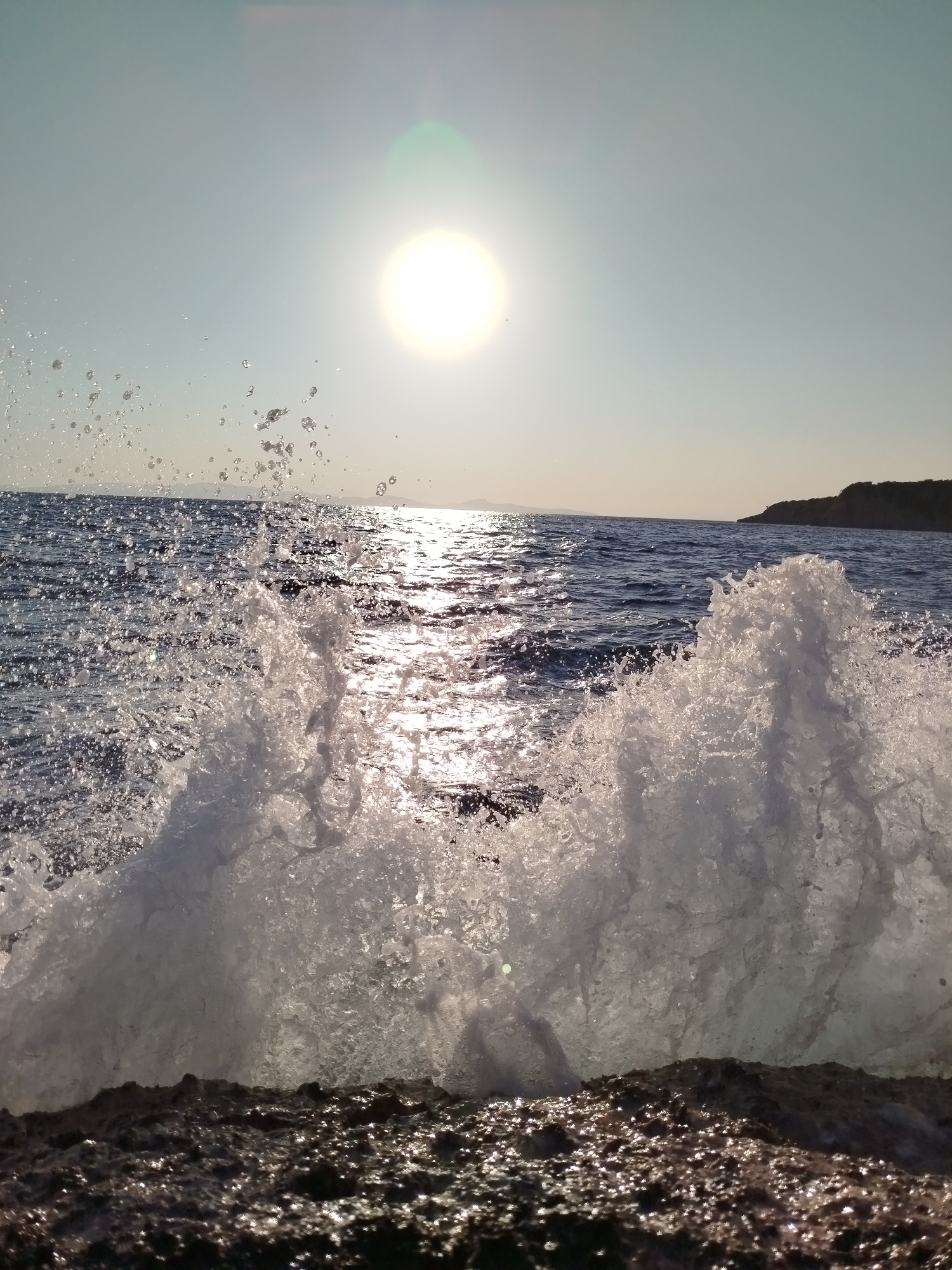A wave crashes against a rocky shore in Karaburun, Turkey. The sun is low in the sky, casting a golden glow on the water and creating a lens flare in the upper center of the image. The wave is captured in mid-air, with water droplets spraying in all directions. The rocky shore is dark and textured, with some areas appearing wet and shiny. The sea is a deep blue, with gentle waves rolling in the distance. A distant shoreline with a dark, hilly landscape is visible on the right. The image is taken from a low angle, emphasizing the power and drama of the crashing wave.