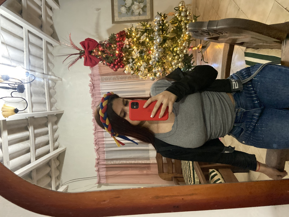 A woman in jeans, a gray t-shirt, and a black jacket stands in front of a mirror taking a selfie with a red phone. She's wearing a colorful braided headband. Reflected in the mirror are parts of a room: a white ceiling with a chandelier, a Christmas tree decorated with lights and ornaments, and a curtained window. A wooden chair and part of a wooden table are also visible.