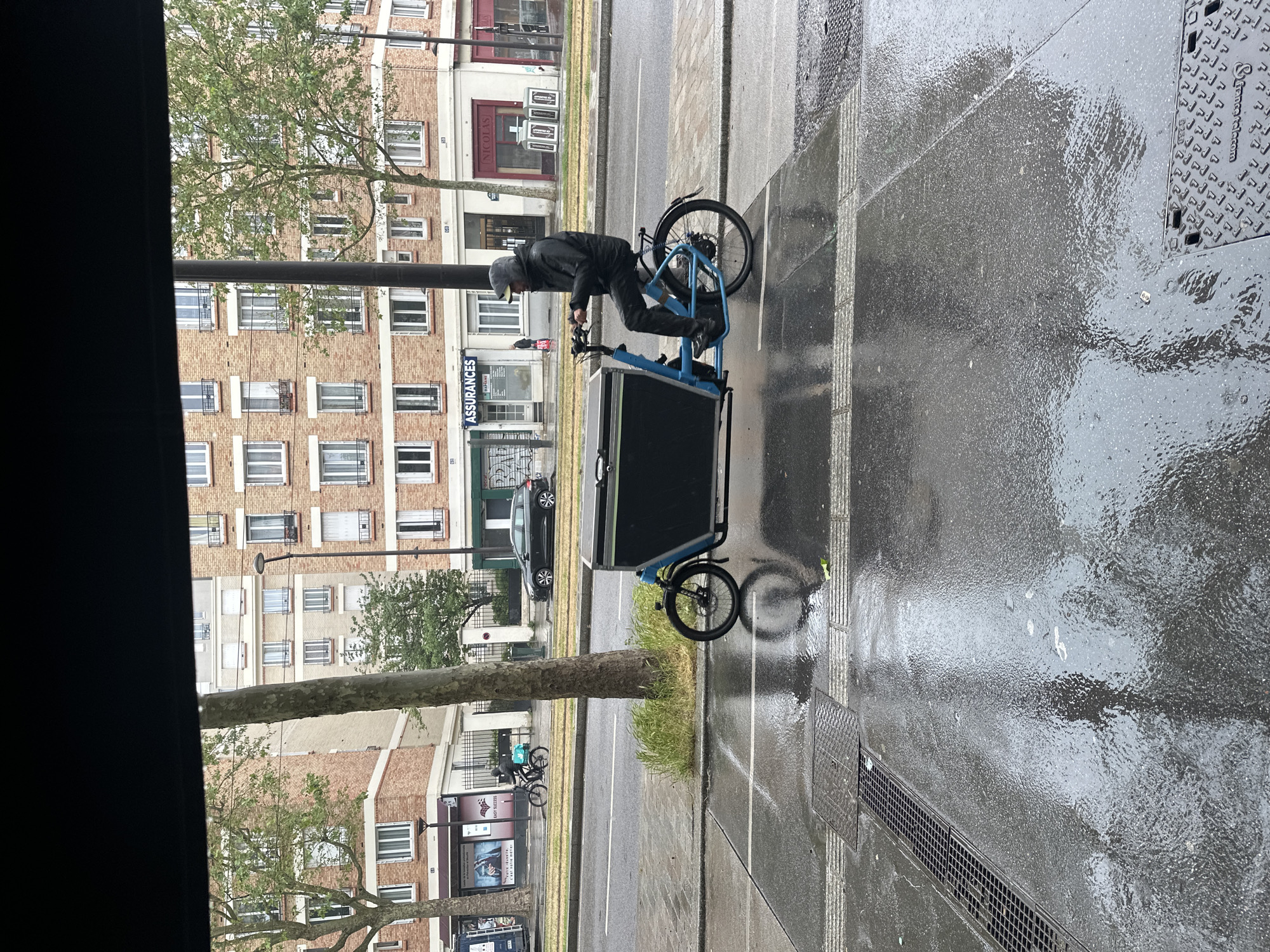 A street-level shot captures a courier riding a blue cargo bike on a wet street in Paris, France. The courier, clad in dark clothing and a cap, is actively cycling. The cargo bike features a sizable black cargo box in front. The street is slick with rain, and reflections are visible on the pavement. Buildings line the street, with the architecture typical of Parisian urban settings. The image is taken from an elevated position, possibly from a window. The scene is urban, with a focus on transportation and delivery services.