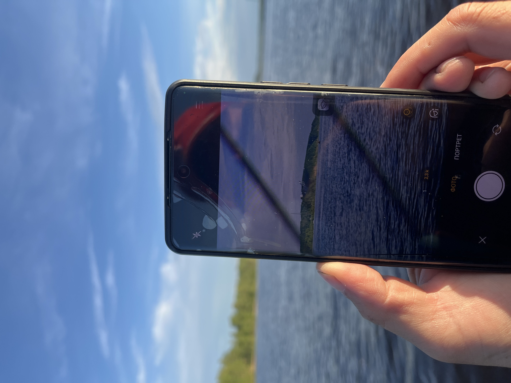 A person's hand holds a smartphone, capturing an image of a wide body of water and a distant tree-lined shore. The smartphone screen displays a landscape scene with choppy water and a hazy horizon, showing icons for camera settings like zoom and modes. The sky above is a bright blue with scattered white clouds, suggesting daylight. The body of water is likely a reservoir, as Chaikovsky, Russia, is known for the Kama Reservoir on the Kama River. The image captures a serene moment, possibly a tourist or local enjoying the scenery.
