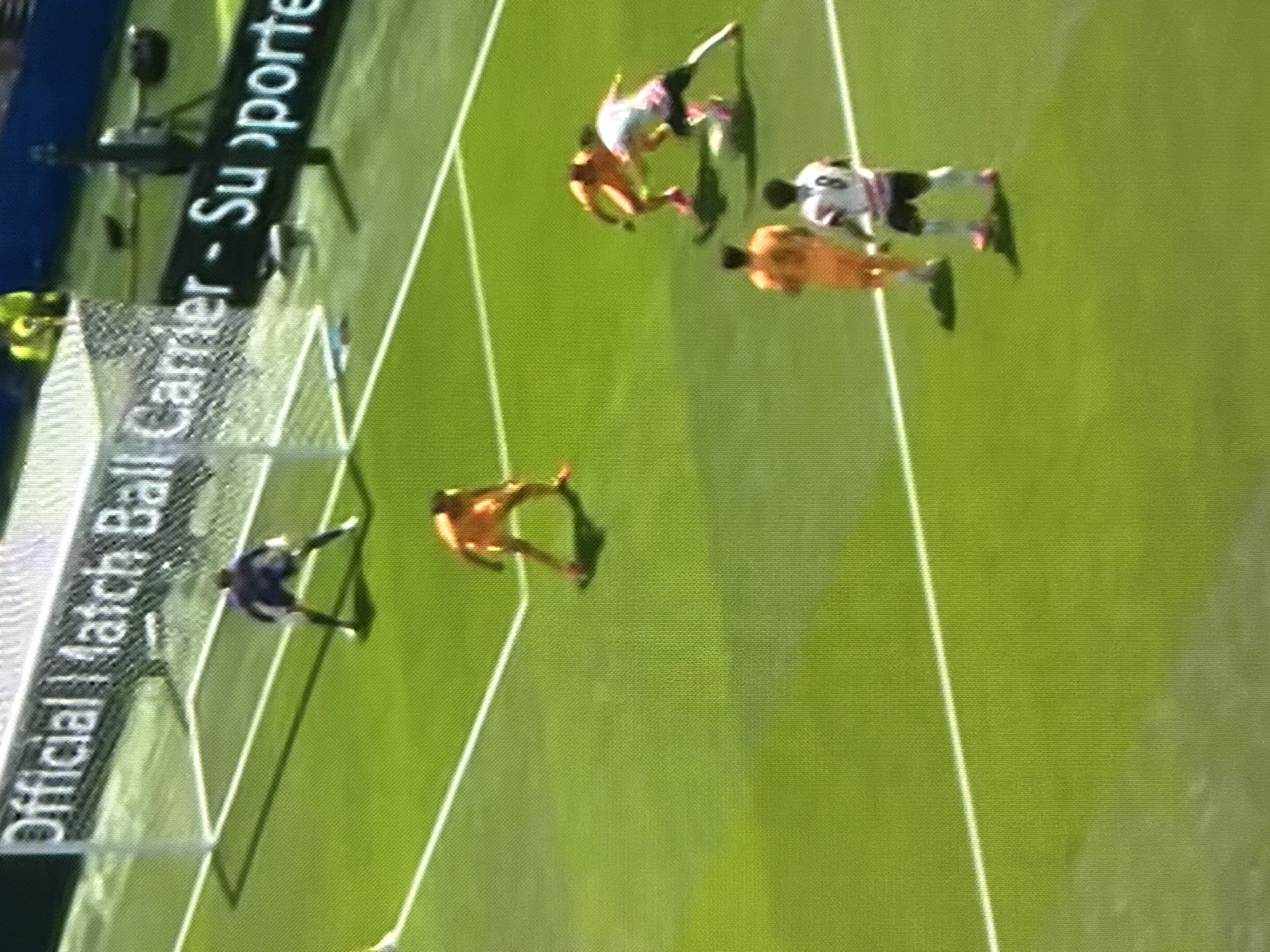 The media file shows a soccer match in progress. There are several players on a bright green field, some in action and others standing. The players are wearing different colored uniforms. One player in a white and black jersey is jumping, with another player in an orange jersey nearby. Another player in a dark blue jersey is in the background near a net. The scene appears to be outdoors on a sunny day, with clear shadows on the field. In the background, there is a banner with white text on a black background. The text is partially visible and reads "Official Match Ball Carrier - Su porte". The overall impression is of a professional sporting event.
