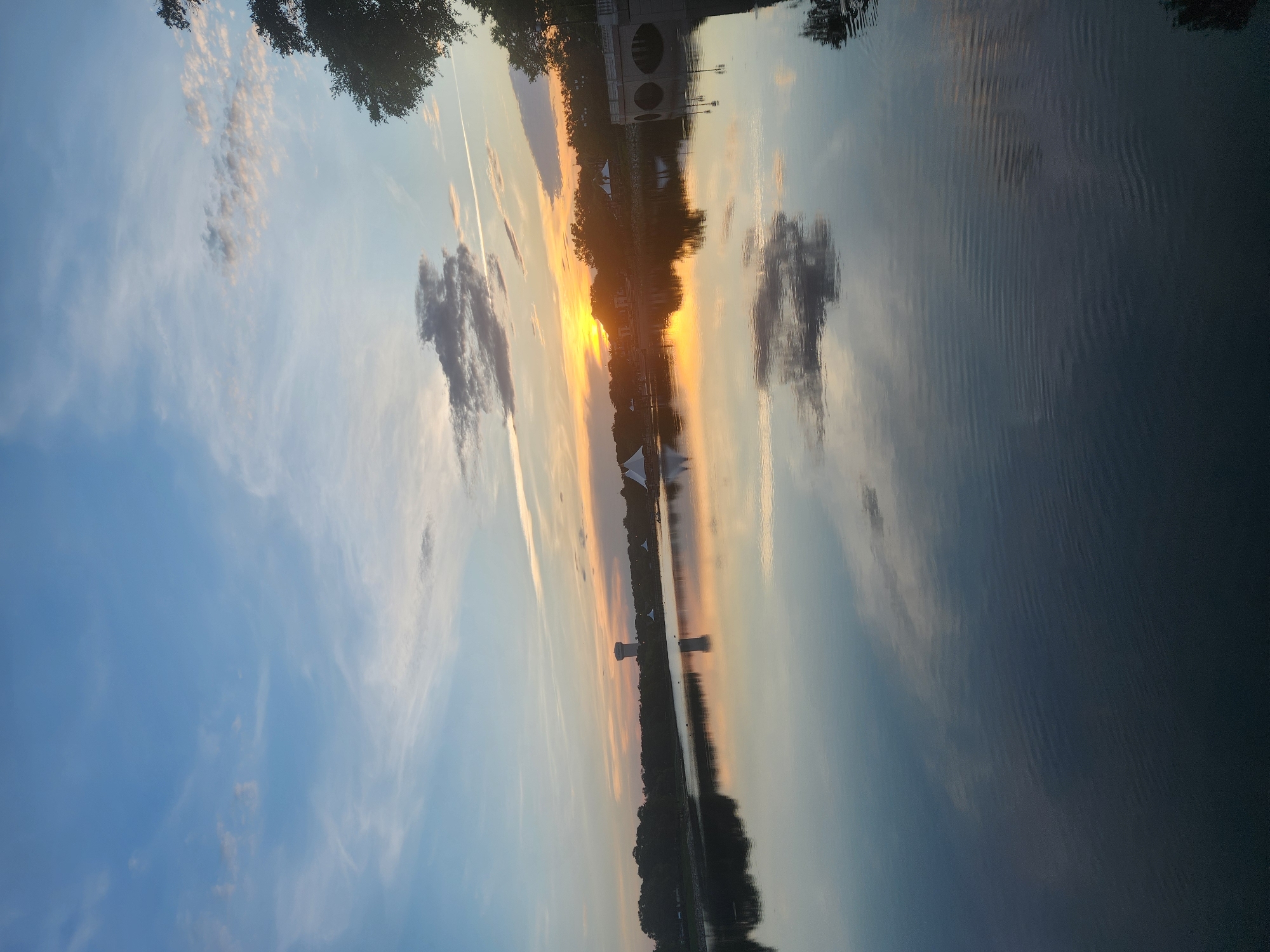 The image depicts a sunset over a calm body of water, likely Lake Eola Park in Orlando, Florida, given the presence of a water tower consistent with those found in the area and the characteristic cityscape visible across the lake. The sky is a gradient of blues and oranges, with wispy clouds scattered across it. The setting sun casts a warm glow on the horizon, reflecting vibrantly on the water's surface. Trees and buildings line the distant shore, and a bridge with arched supports is visible on the right side of the frame. The water itself is still, creating a mirror-like reflection of the sky and the surrounding landscape. There are no people or active events clearly depicted in the scene.