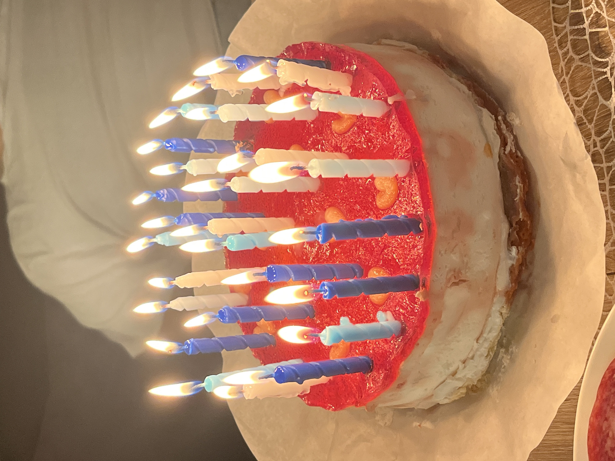 A tilted overhead close-up shows a partially eaten, two-layer birthday cake on parchment paper. The cake has white frosting and is topped with a layer of shiny red gelatin studded with orange slices. Approximately 26 white and blue spiral birthday candles are lit and stuck into the gelatin. A person wearing a gray shirt stands beside the cake. A wooden table with a mesh covering is visible to the right, along with the edge of a white plate holding round, red food slices. The image location is not identifiable as Saarbrücken, Germany.